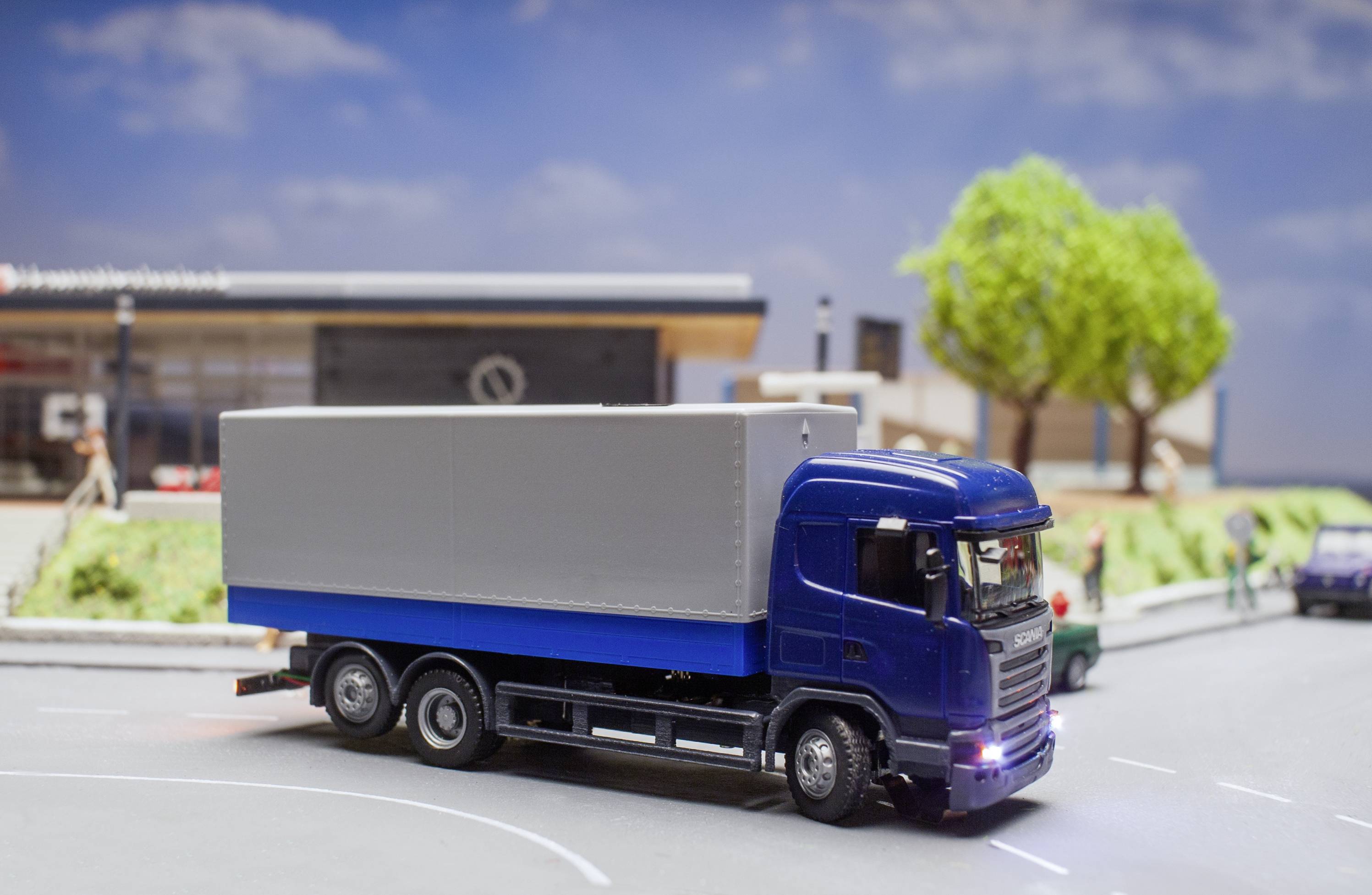 A blue lorry drives along a model road in front of a miniature backdrop with trees and buildings in the background.