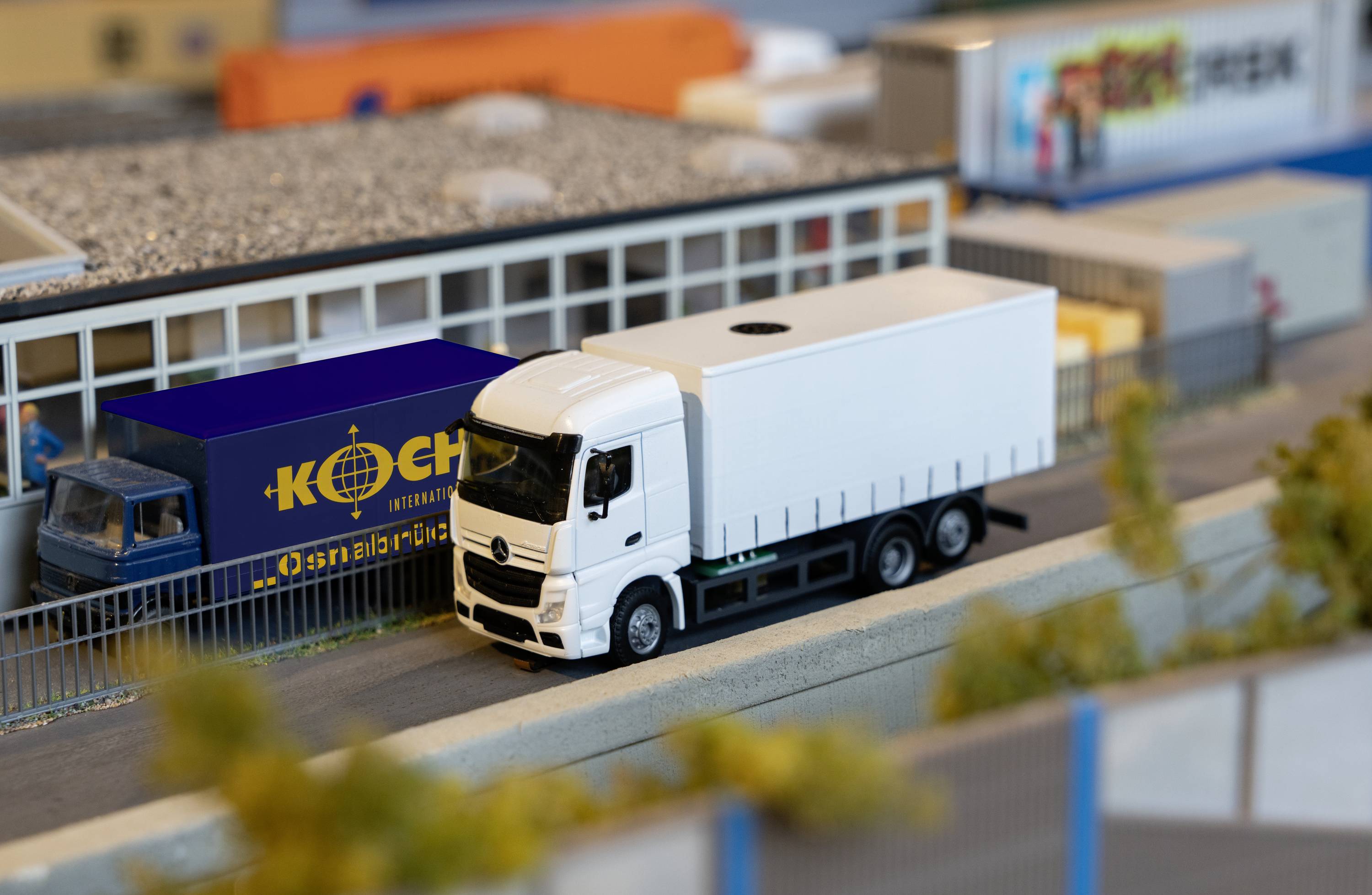 A white lorry is driving on a model road next to a company building. Shipping containers are visible in the background.