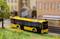 A yellow model bus travels along a miniature road beside buildings, a tree, and a green area.