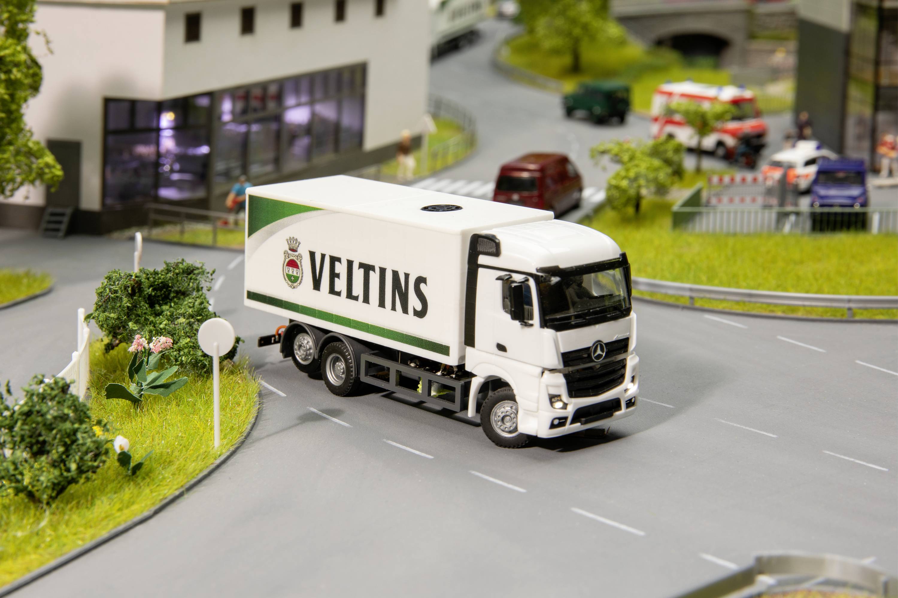A white lorry with 'Veltins' written on its side is driving along a modelled street landscape. Buildings and trees can be seen in the background.