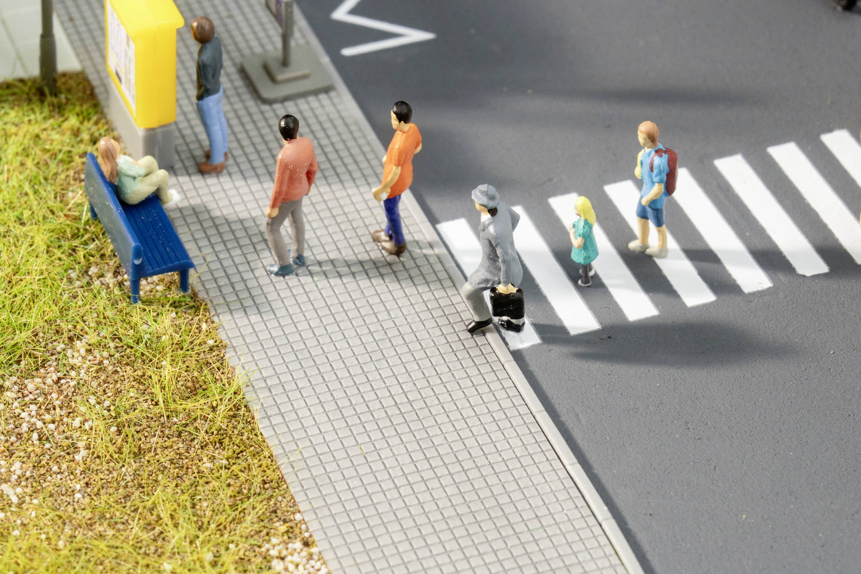 Toy figures are crossing a zebra crossing on a miniature street. A person is sitting on a bench. Scenario of an urban environment.