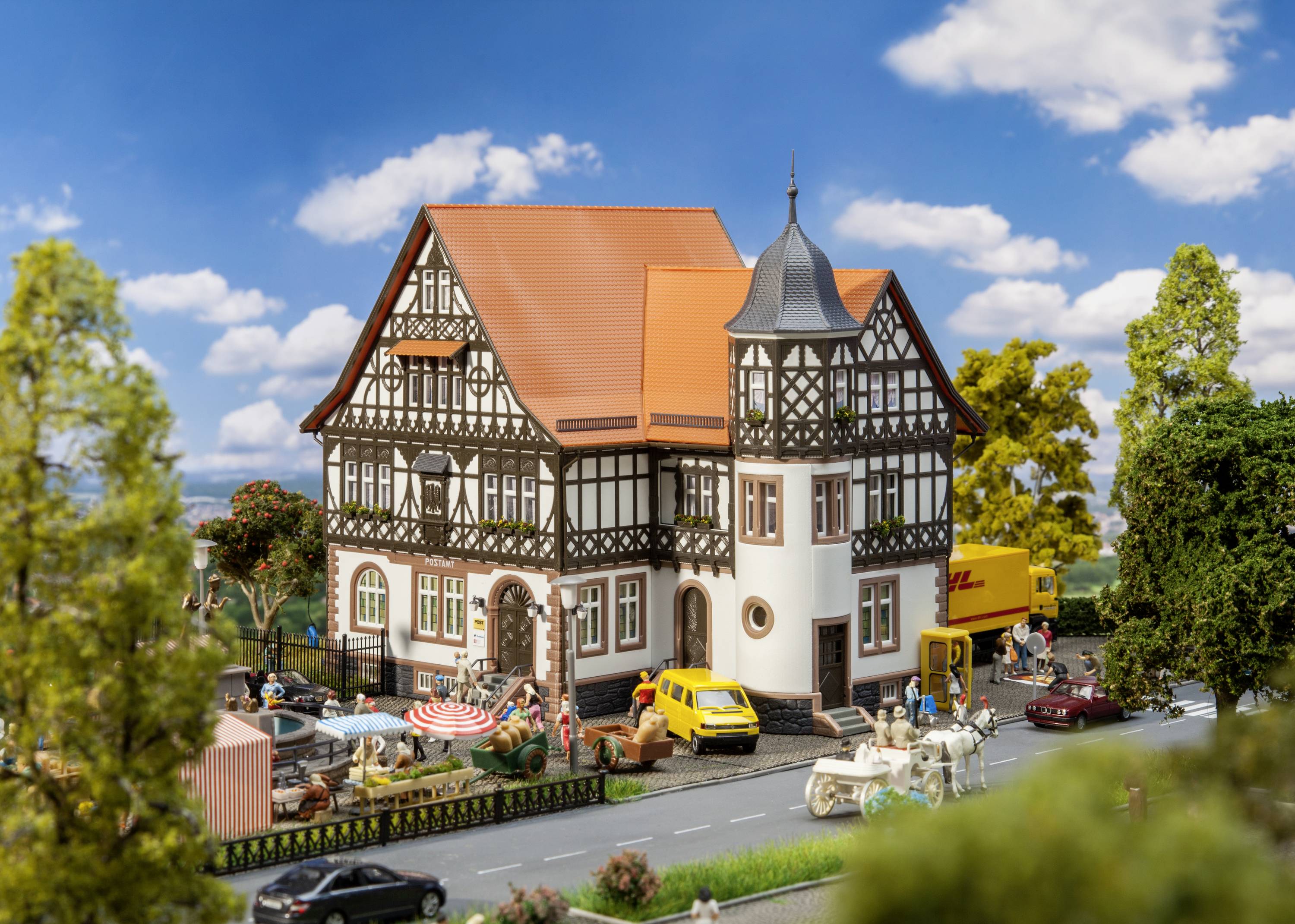 A model railway landscape depicts a historic half-timbered house with an orange roof. People, cars and trees are visible in the foreground.