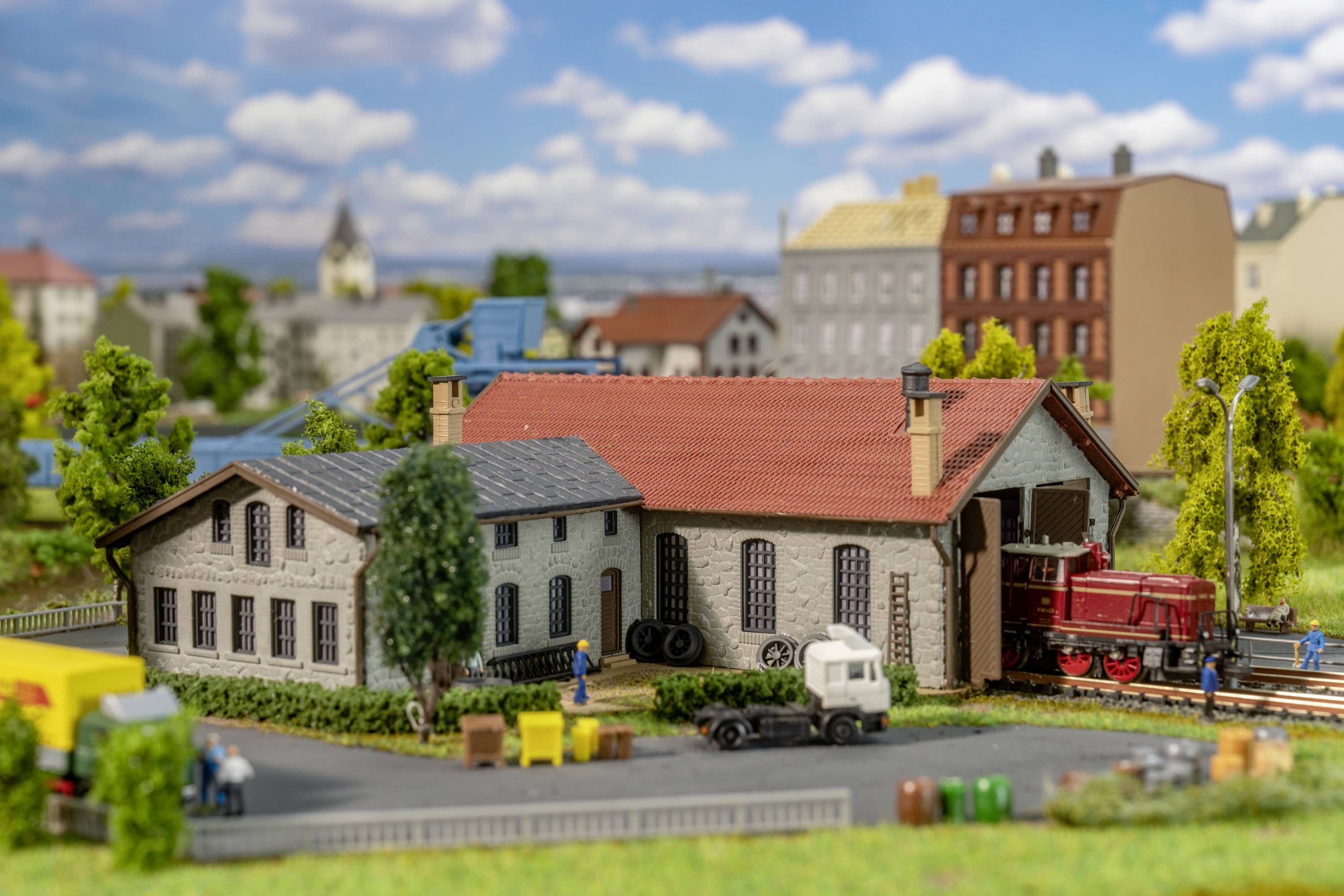 A model railway layout with a goods station, locomotive, and buildings in a rural setting. People and vehicles can be seen.