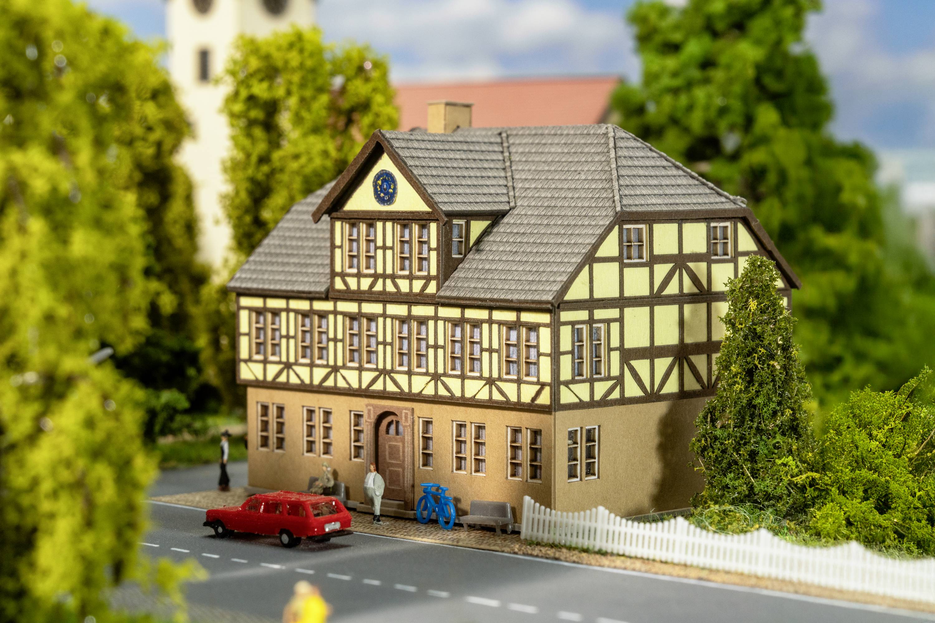 A scale model of a half-timbered house with a red car and blue bicycle in front, surrounded by trees, on a street.
