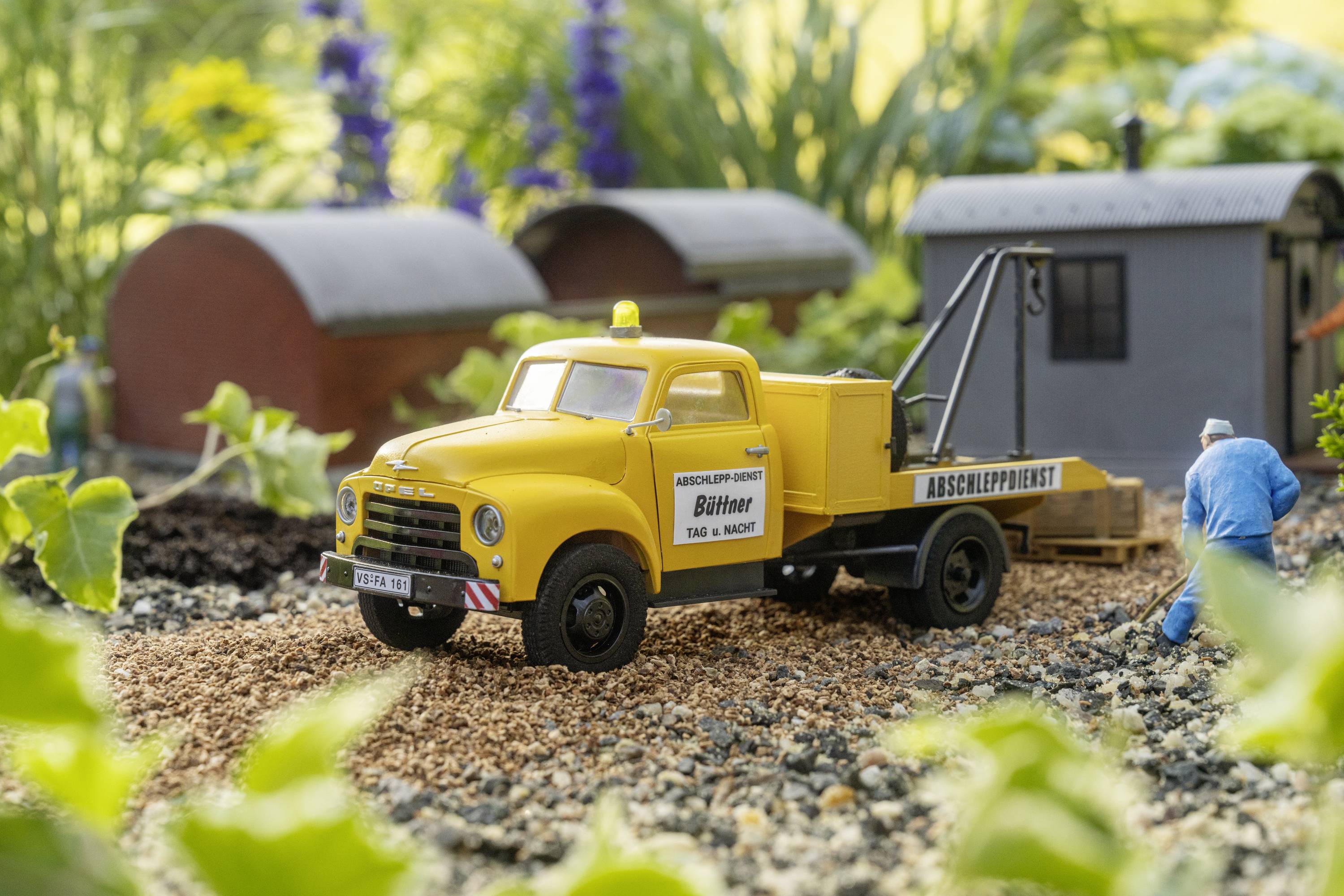Yellow recovery vehicle on a modelled gravel road. Miniature buildings and plants in the background. A model maker is working on details.