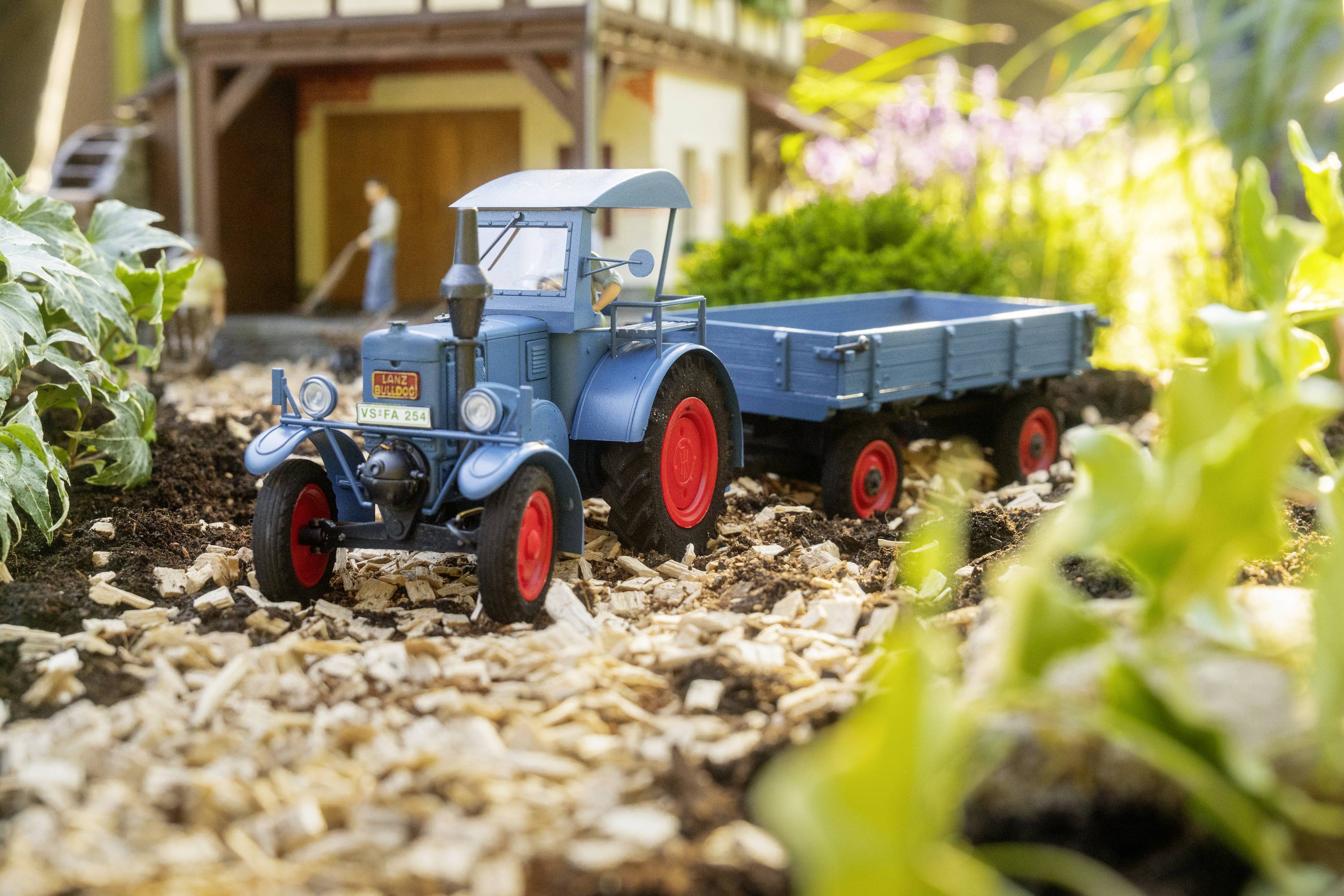 A blue model tractor with a trailer is standing on a garden path, surrounded by plants, with a miniature house in the background.