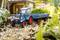 A blue model tractor with a trailer is standing on a garden path, surrounded by plants, with a miniature house in the background.