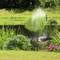 A heron stands on the bank of a pond, beside flowering hydrangeas. In the background, a water sprinkler sprays water into the air.