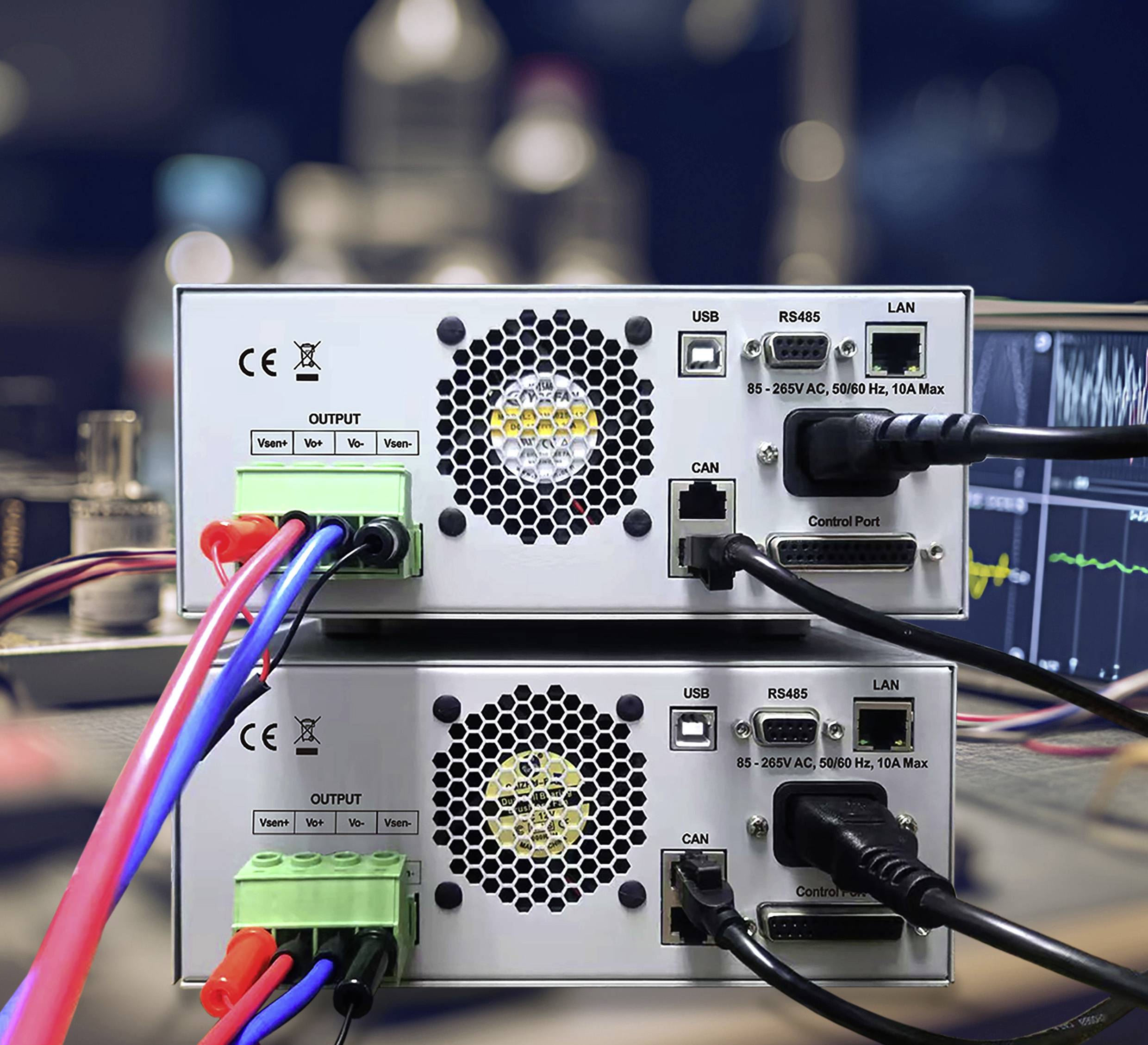 Two electronic devices stacked, featuring USB, RS485, LAN ports, and labeled input/output connections, with cables attached. Background shows a blurred control room.