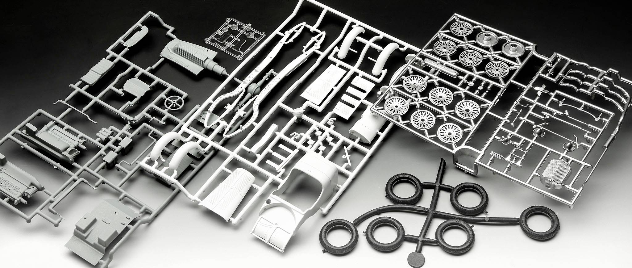 A plastic model kit with various parts, including tyres, body panels and vehicle components, on a grey surface.