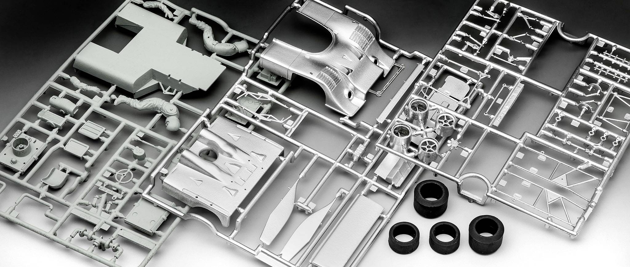 Parts of a plastic model kit in a casting frame, including car body parts and tyres, are laid out in an orderly manner on a table.