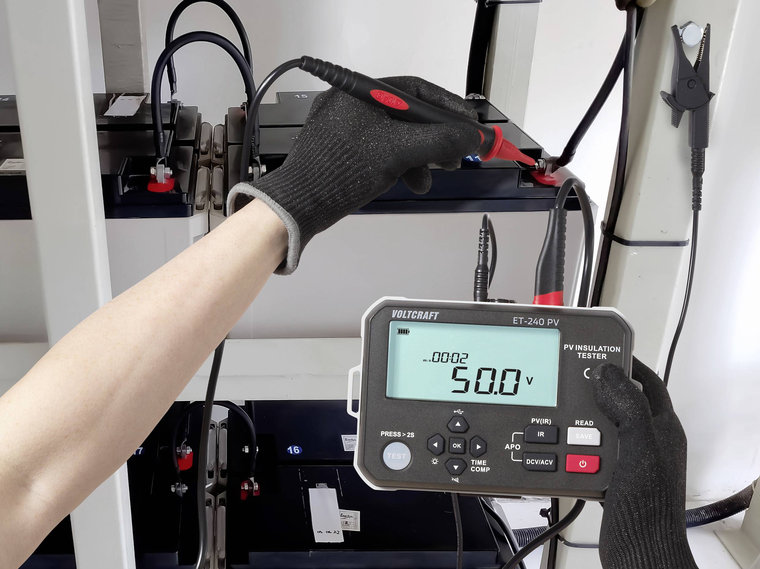 'Person measuring battery voltage with a digital multimeter. The display shows 50.0 volts, indicating the current voltage reading.'