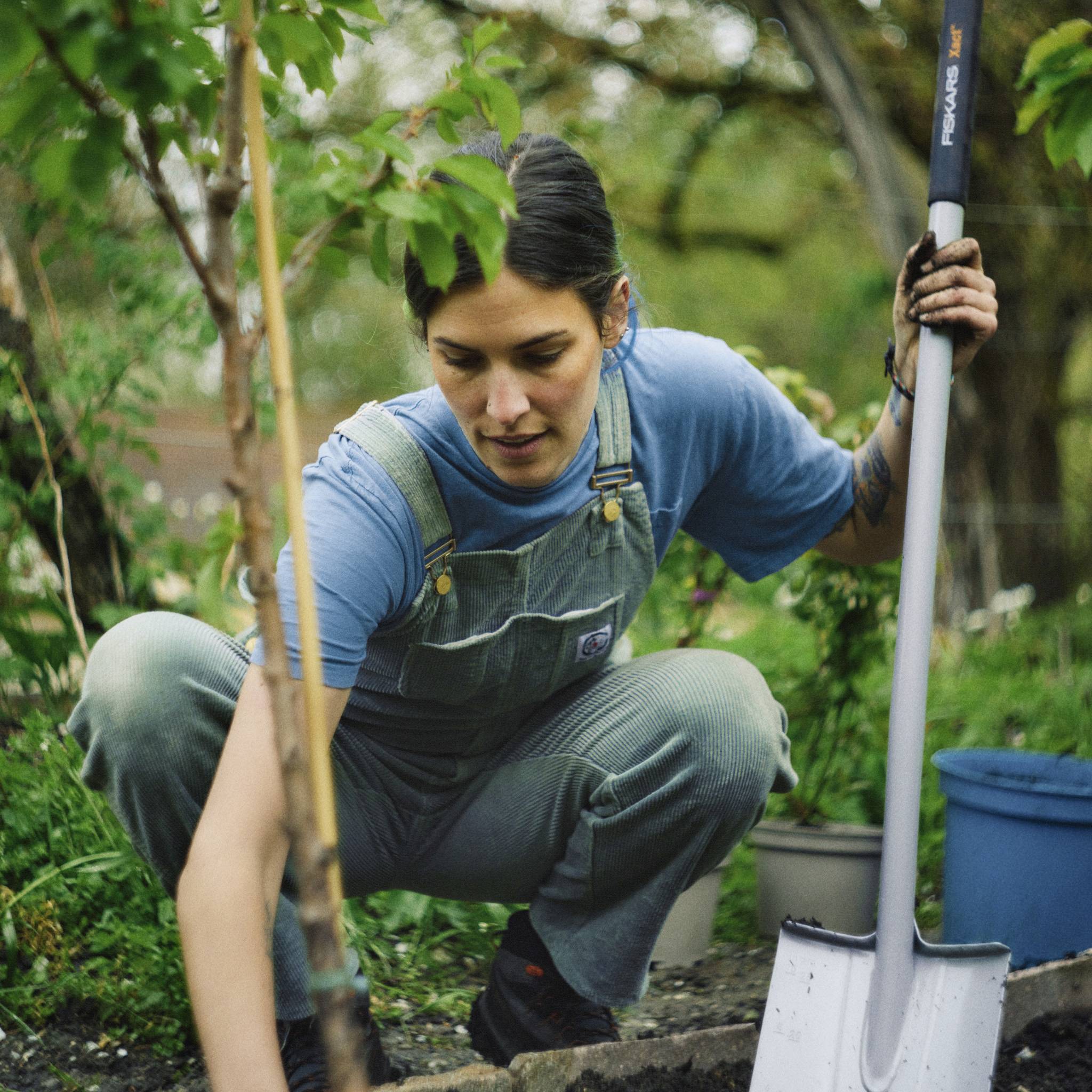 A woman wearing dungarees squats in a garden and works with a spade. In the background, plants and trees are visible.