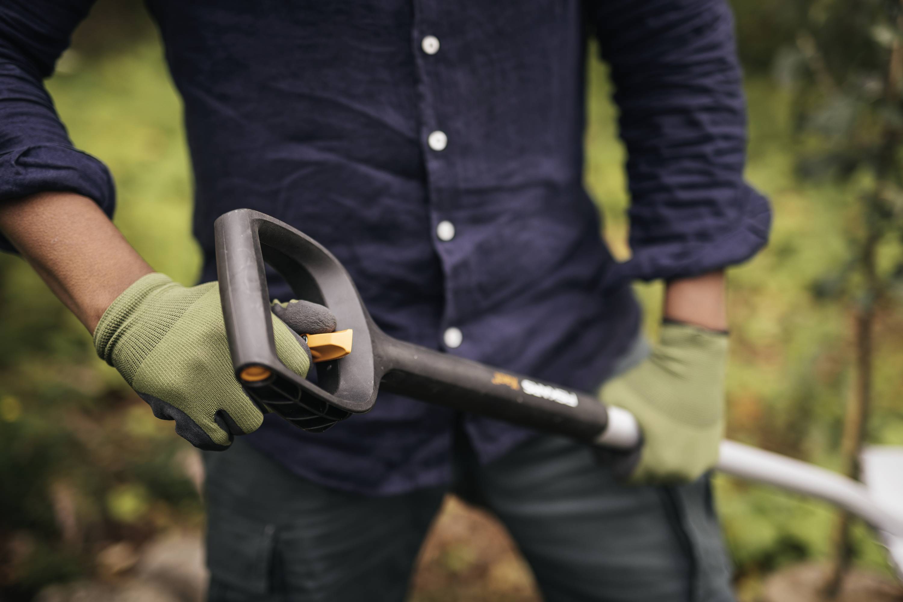 A person wearing a navy blue shirt and green gloves is holding a garden spade. Blurred greenery can be seen in the background.
