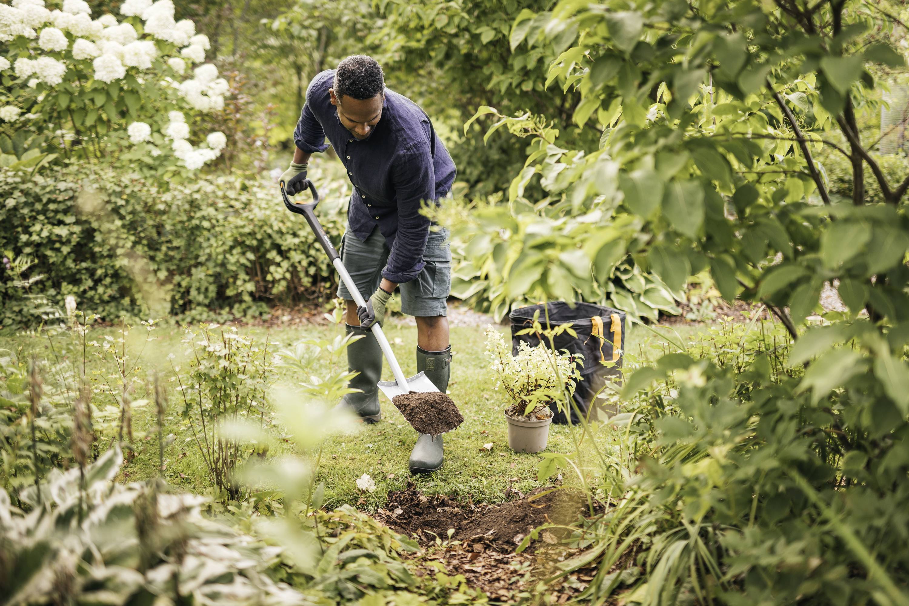 A man is planting in a verdant garden, holding a spade and standing near a small pot with plants. Trees and flowers surround him.
