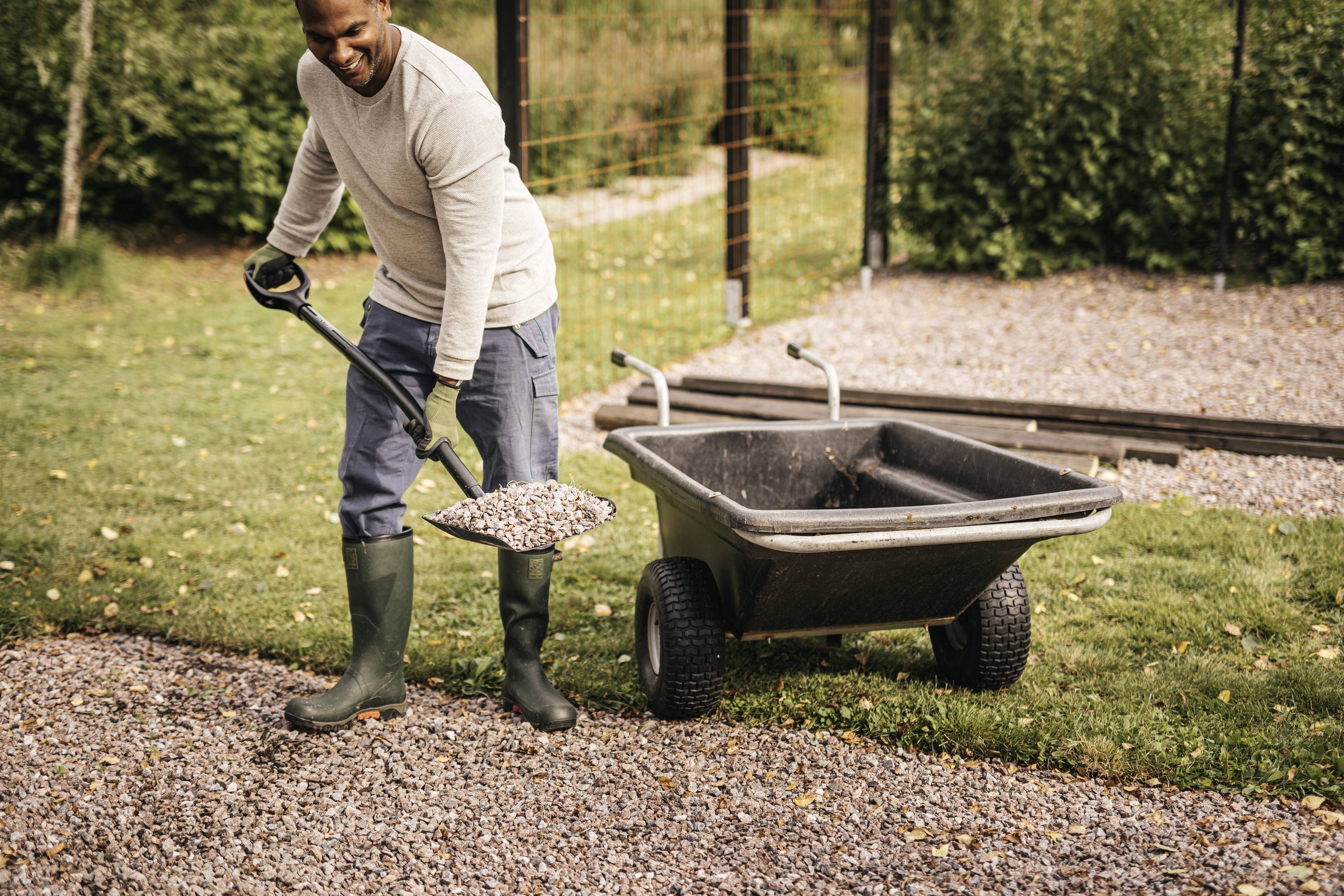 A man in gardening gloves is shovelling gravel onto a path. A wheelbarrow stands next to him. The garden is green and sunny.