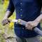 A person is cutting a branch outdoors with an orange-grey, foldable handsaw. The background shows blurred green vegetation.
