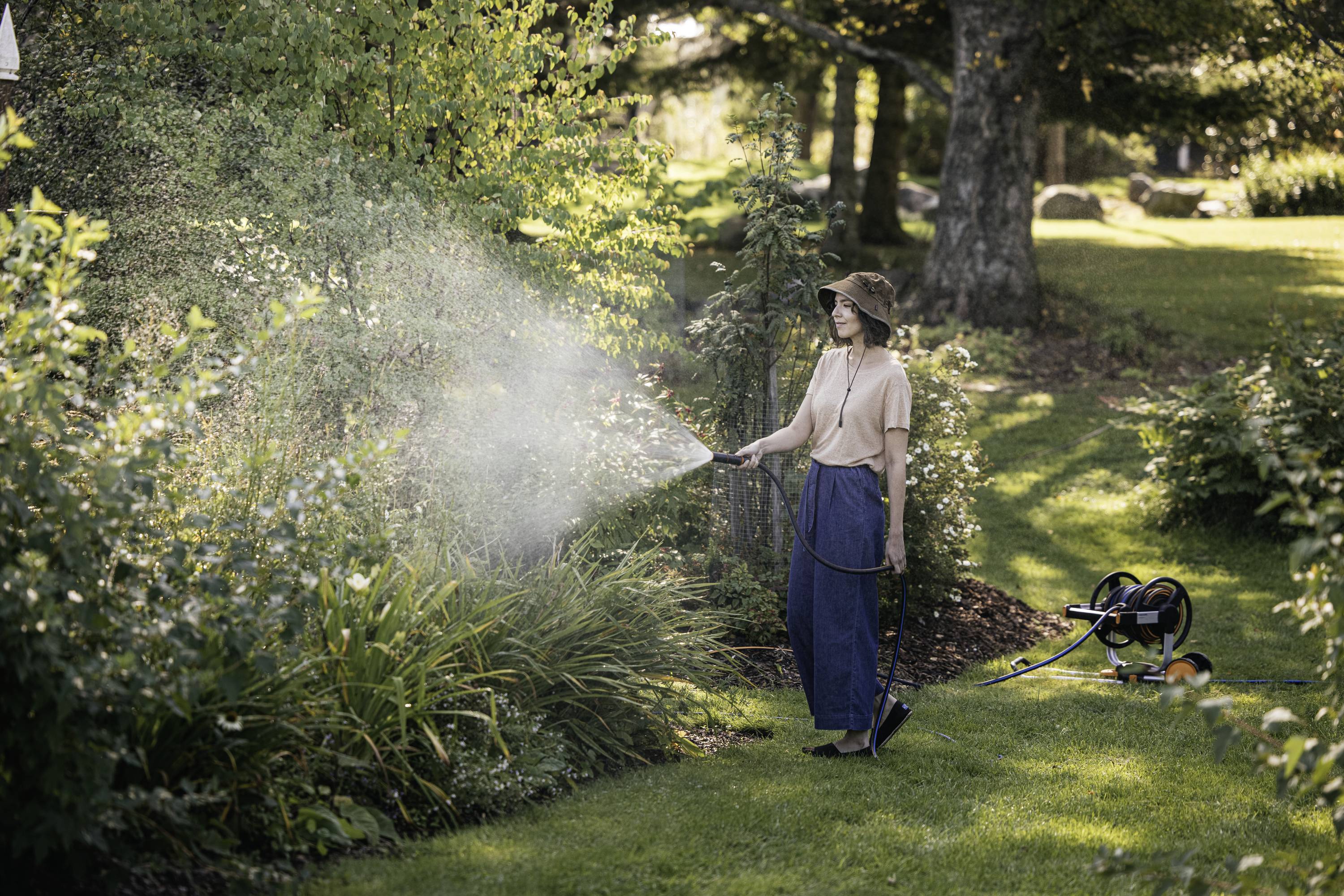 A person is watering plants with a hosepipe in a garden on a sunny day. The surroundings are green and peaceful.