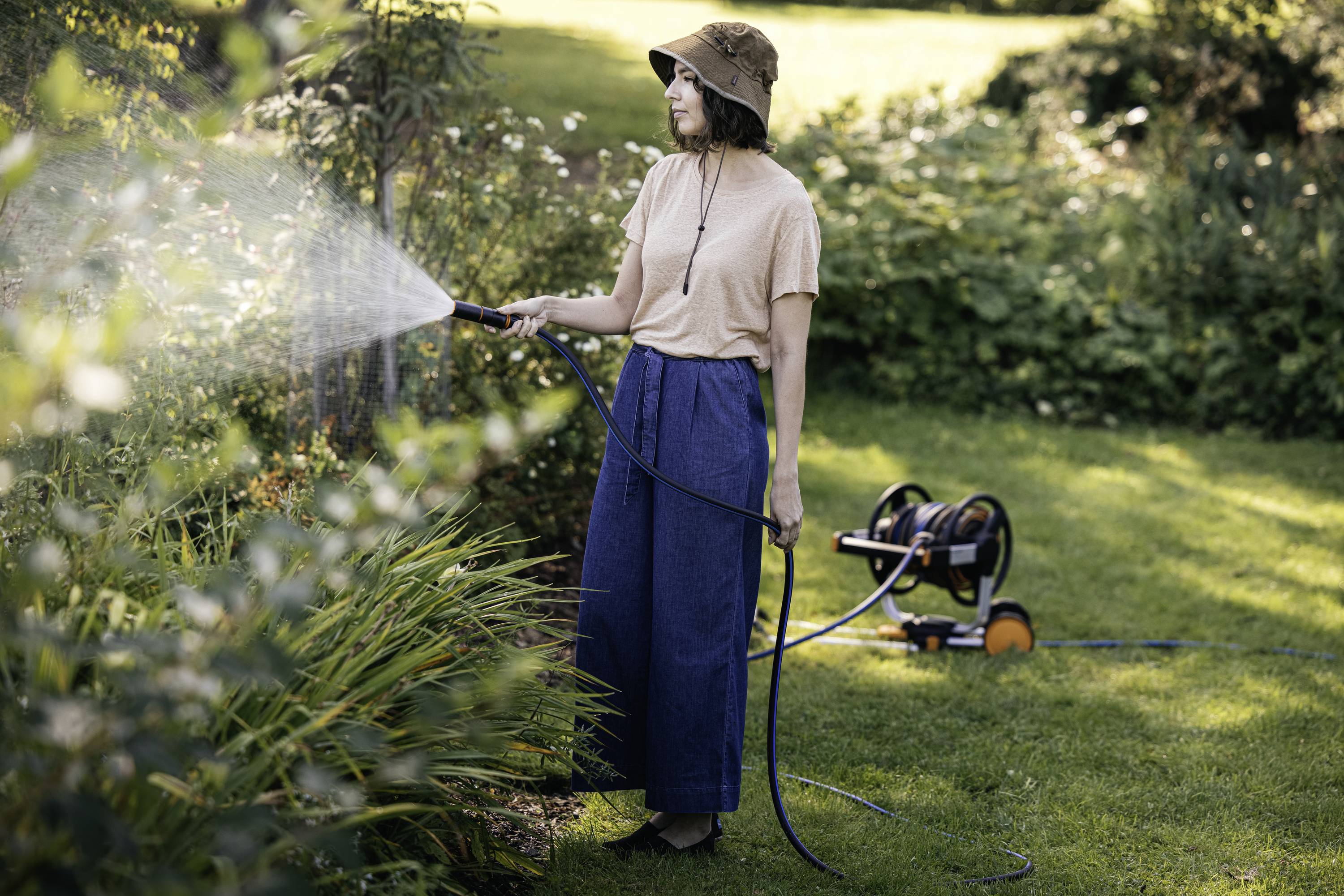 A person wearing a hat is watering plants in the garden with a hosepipe. A hose reel trolley is visible in the background.
