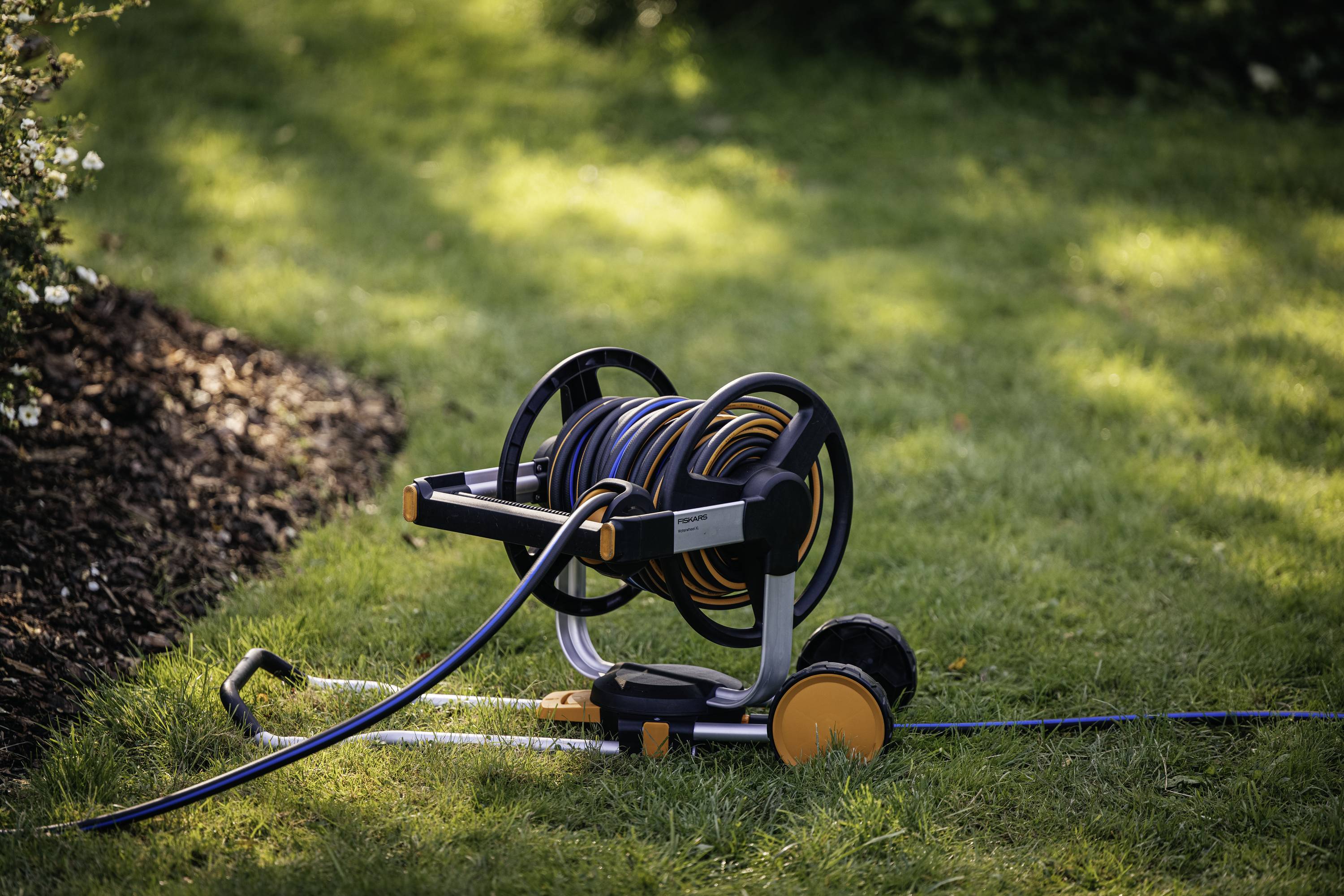 Garden hose on a reel stands on a lawn area, surrounded by green vegetation and bushes in sunlight.