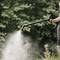 A person is watering plants outdoors with a garden hose. The water stream is strong and the surroundings are lushly green.