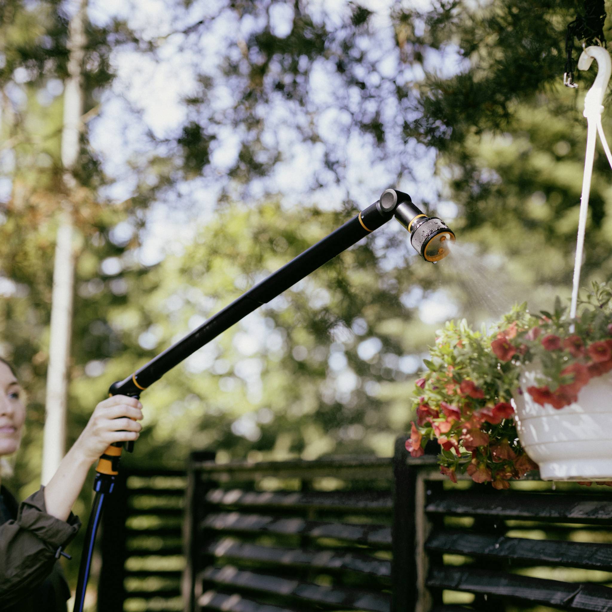 A person is watering hanging flowers with a garden hose. Trees and a wooden fence can be seen in the background.