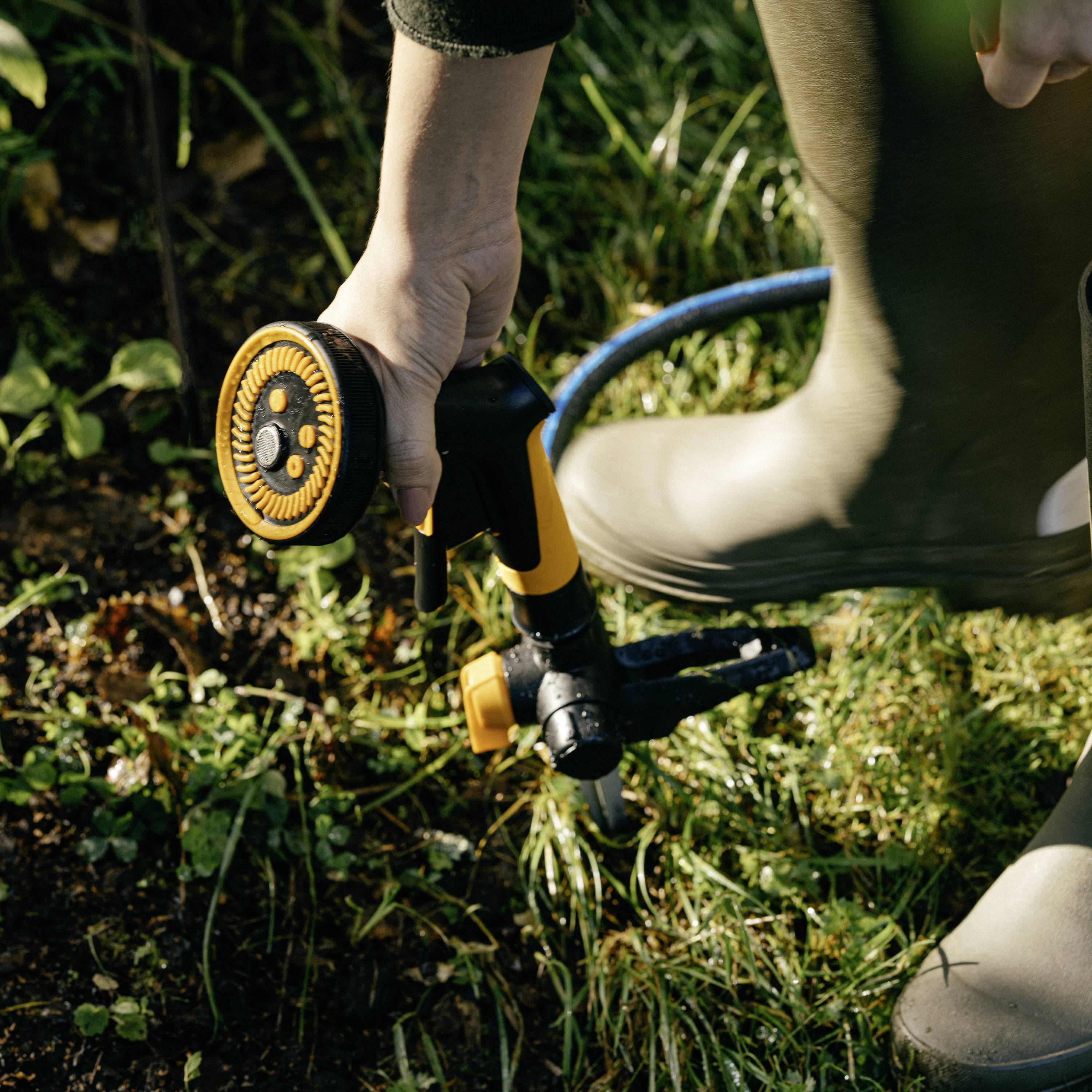 A person is watering plants with a garden hose. Sunlight illuminates the green grass in the background.