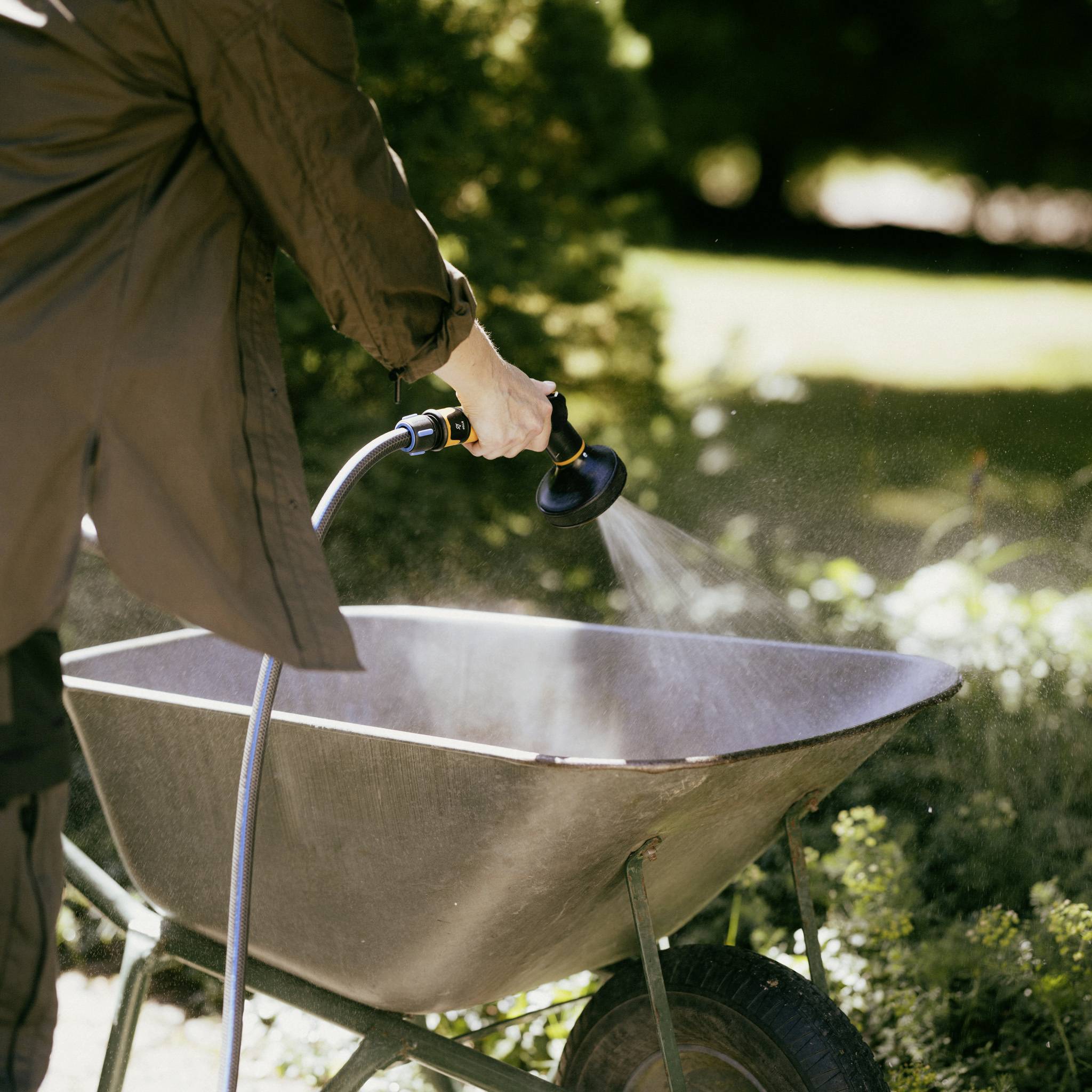 A person is watering a wheelbarrow with a hosepipe. The scene takes place outdoors, surrounded by green grass.