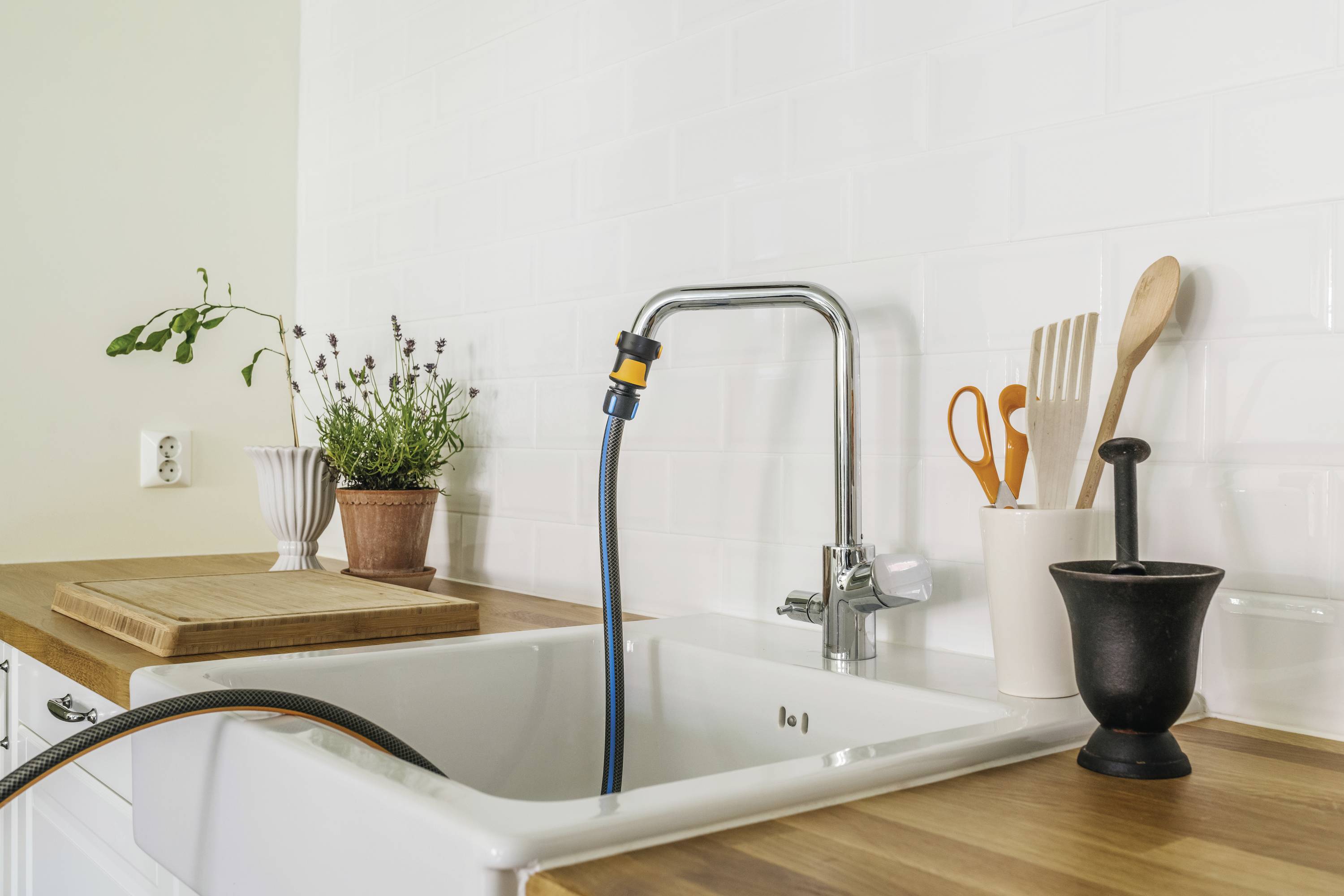 A kitchen sink with a mix of wooden and ceramic utensils. A tap with an attached hose, plants in the background.