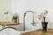Kitchen worktop with sink, chopping board, plants, kitchen utensils in a container and a pestle against a white tiled wall.