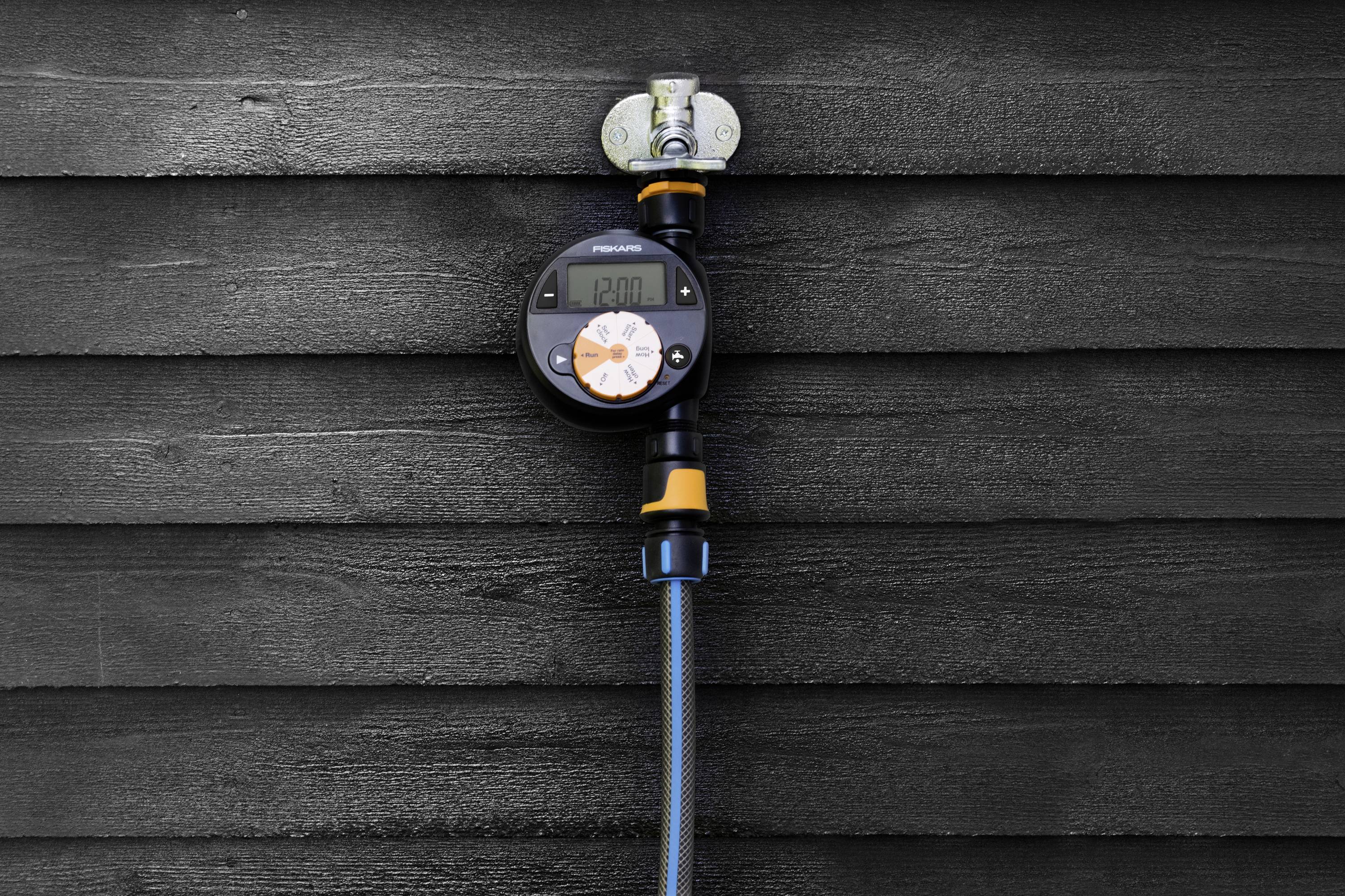 A digital irrigation timer is mounted on an exterior wall, displays the time, and is connected to a blue garden hose.