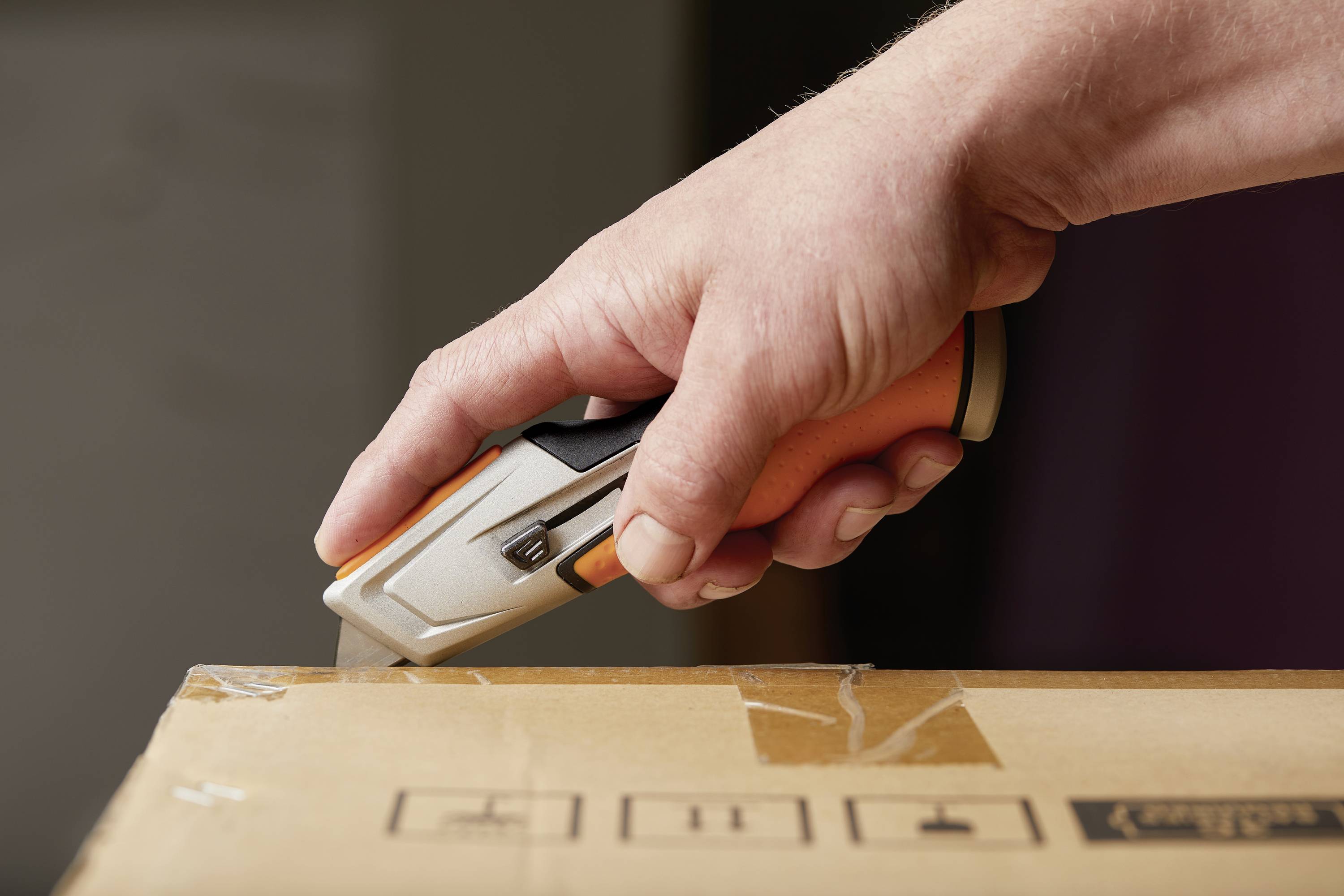 A hand holds a craft knife and cuts adhesive tape on a cardboard box.