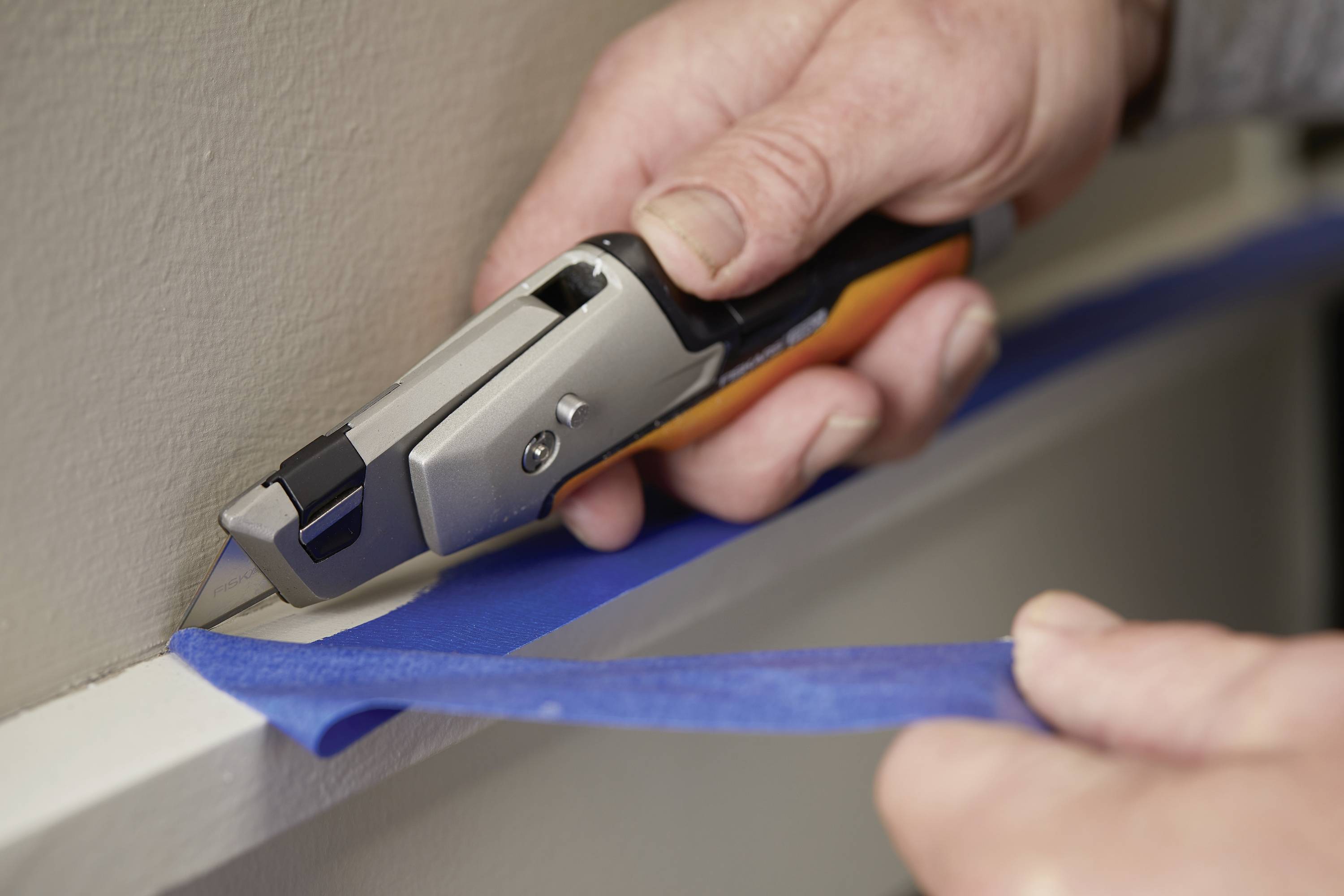 A person is cutting a strip of blue masking tape along the edge of a wall using a utility knife.