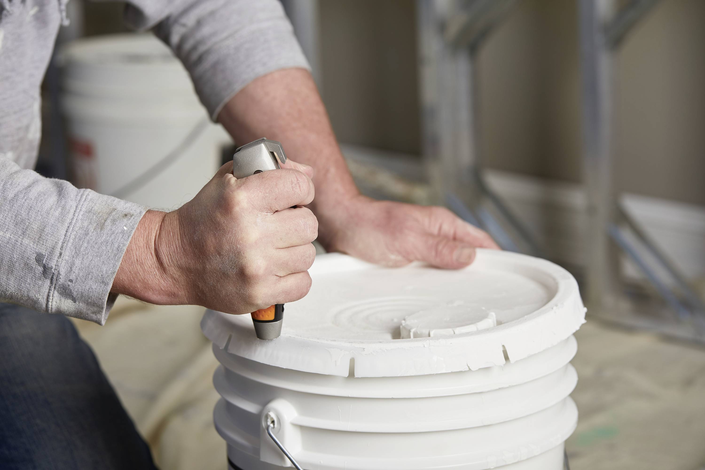 A person is opening a white plastic bucket with a tool. Additional buckets and a shelf are visible in the background.