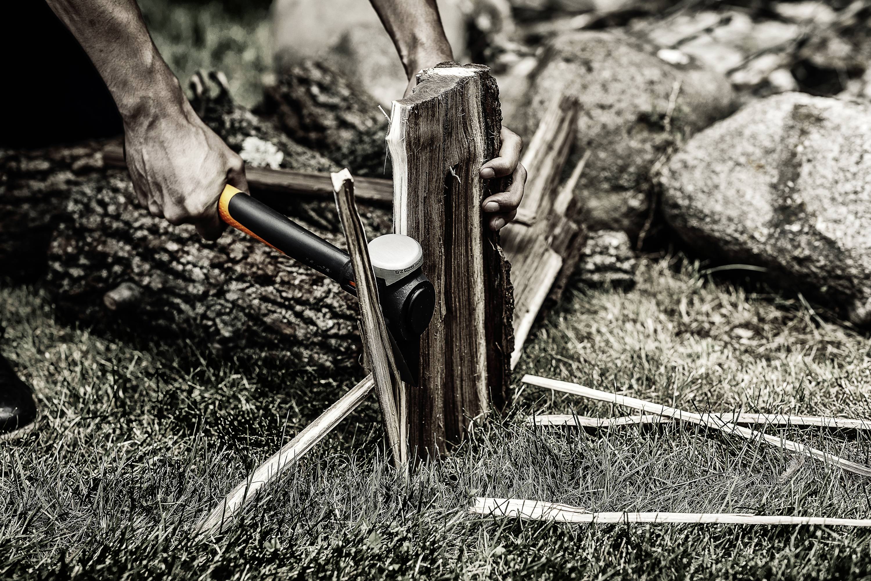 A person is chopping a piece of wood with an axe outdoors. The wood is lying on grass, surrounded by tree trunks and stones.