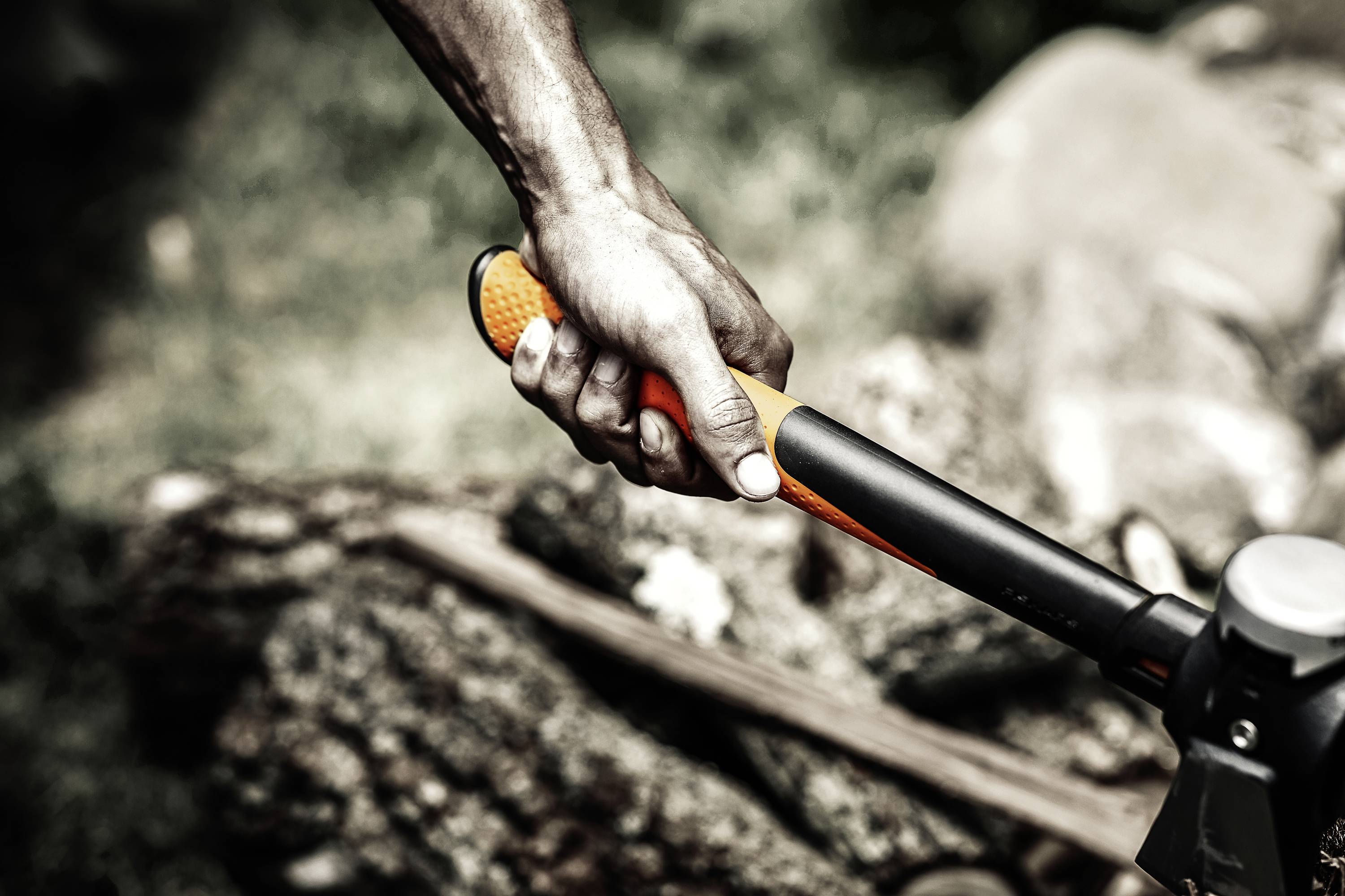 A hand grips the handle of an axe over a pile of wood, poised to split it. The background shows blurred wood and grass.