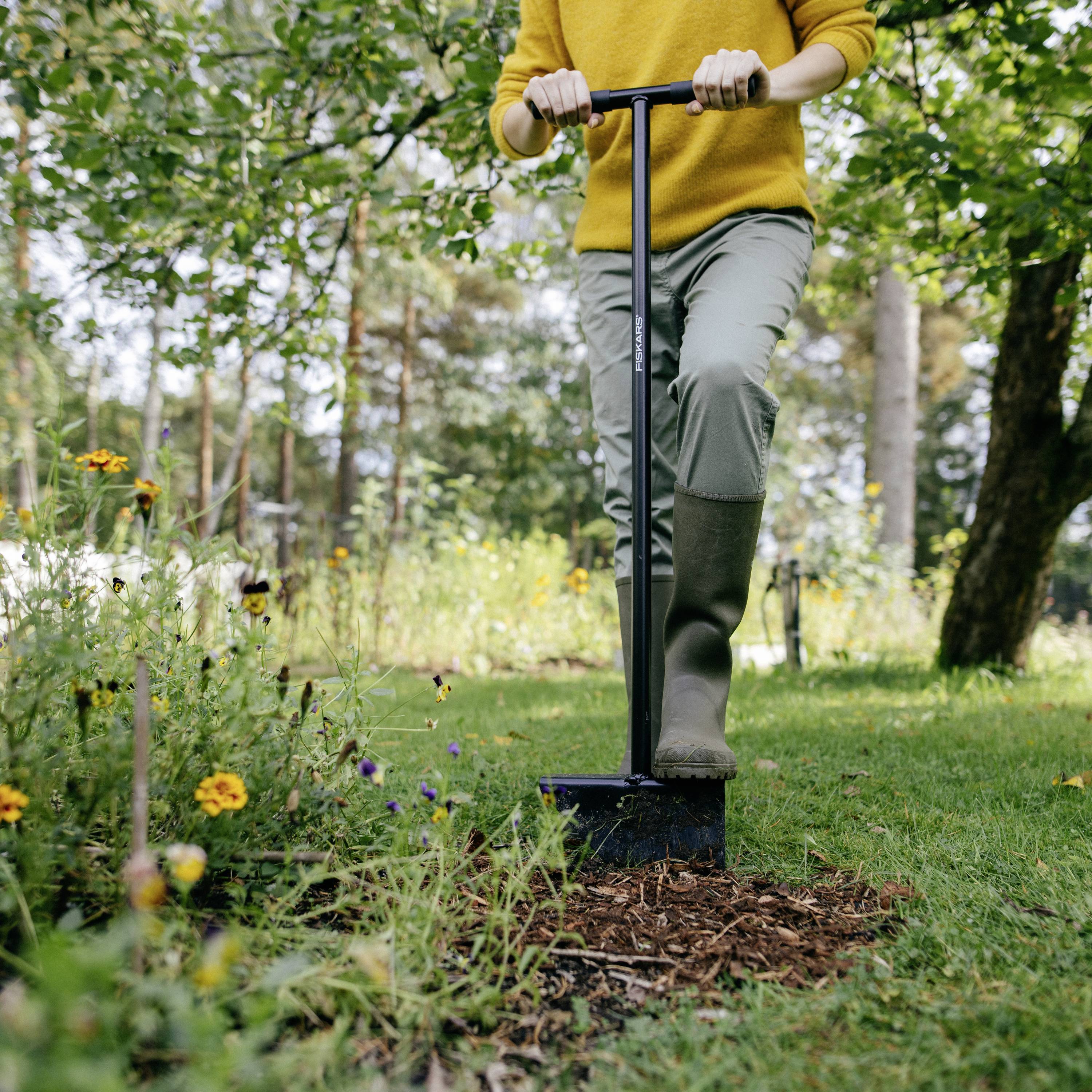 A person is digging with a spade in a garden, surrounded by flowers and trees. They are wearing a yellow top and wellington boots.