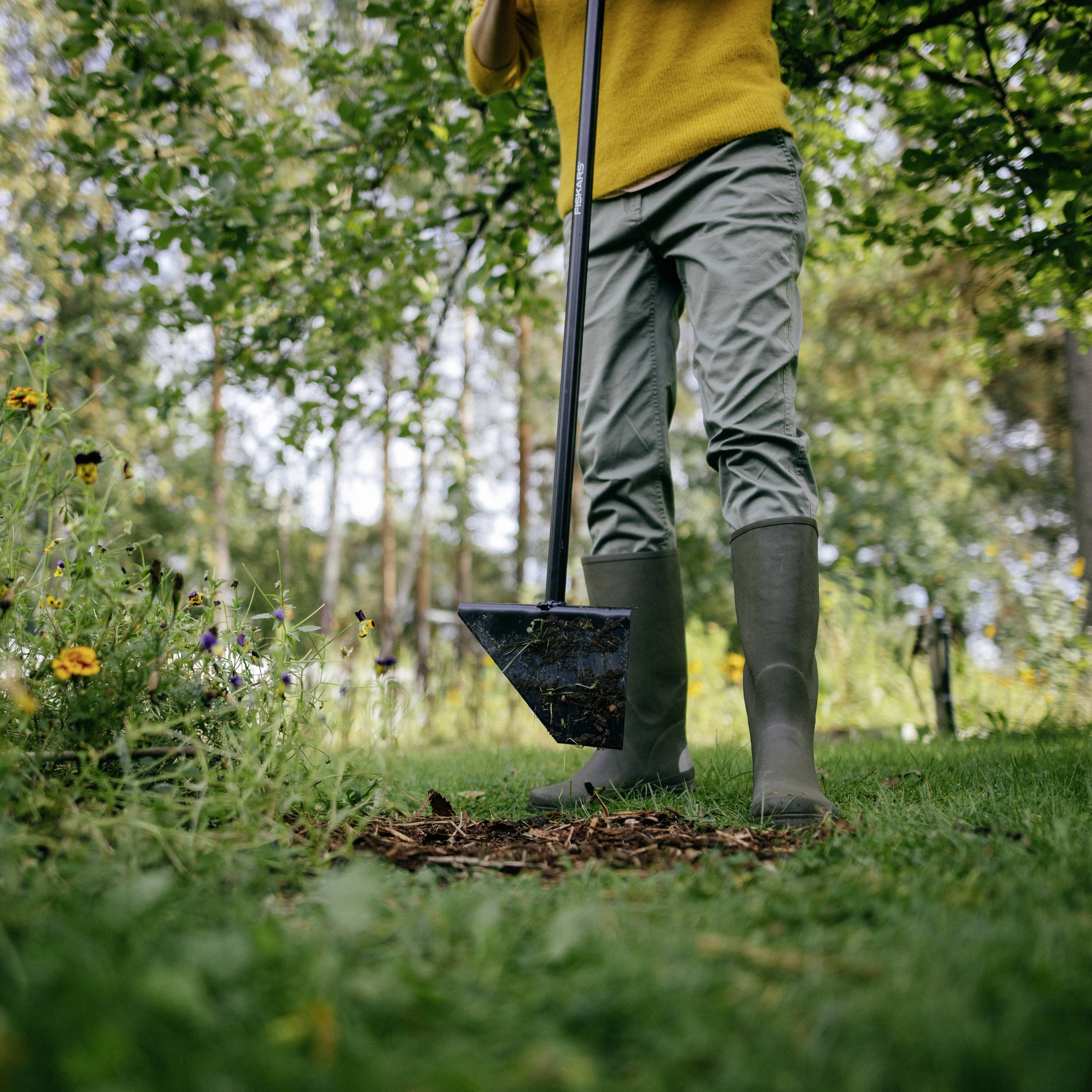 A person is digging a hole with a spade in a flowering garden. They are wearing wellington boots and standing on a green lawn.