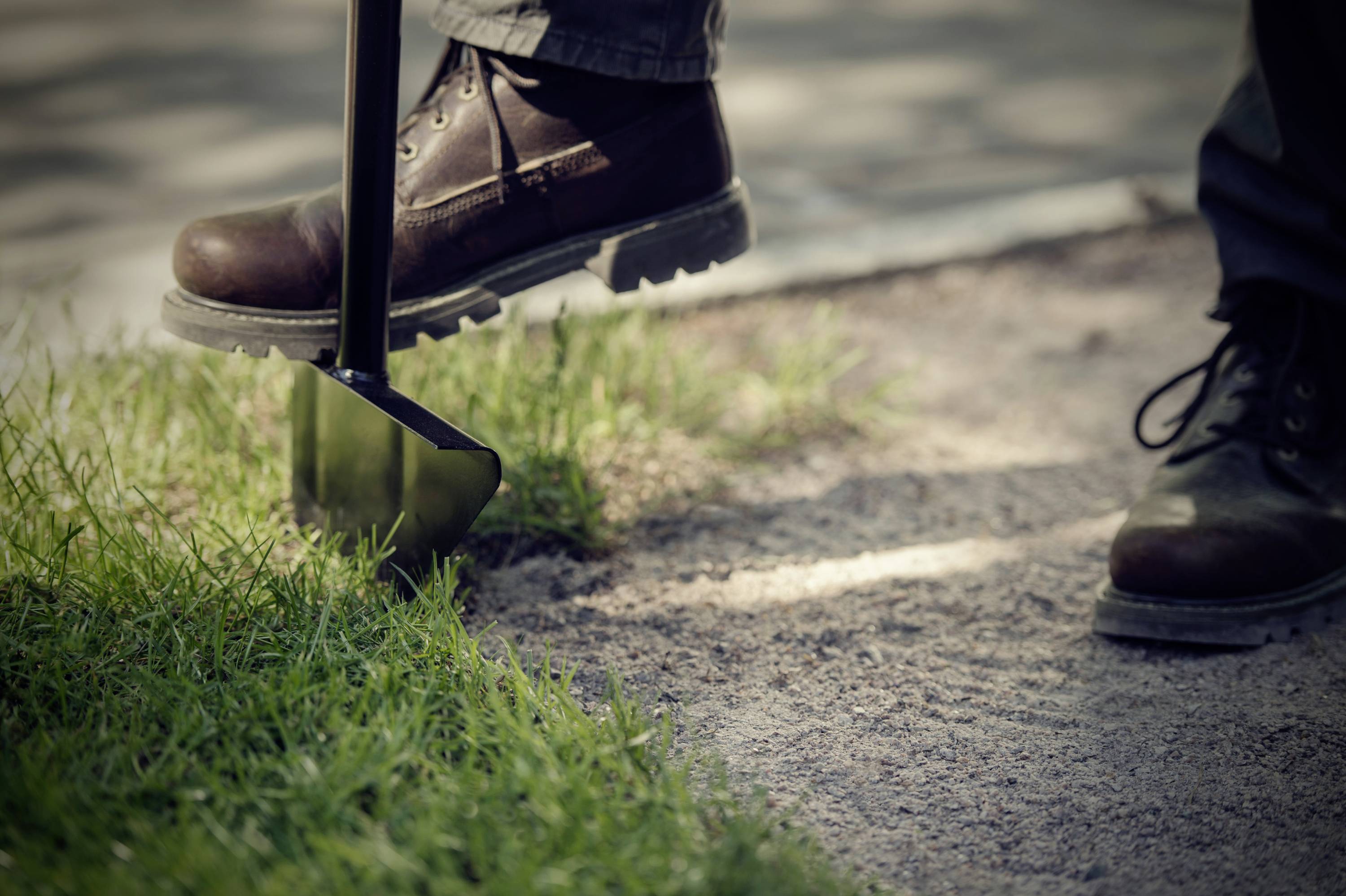 Two people are wearing heavy boots. One is lifting soil with a spade at the kerb's edge. Outdoor work in the garden or landscaping.