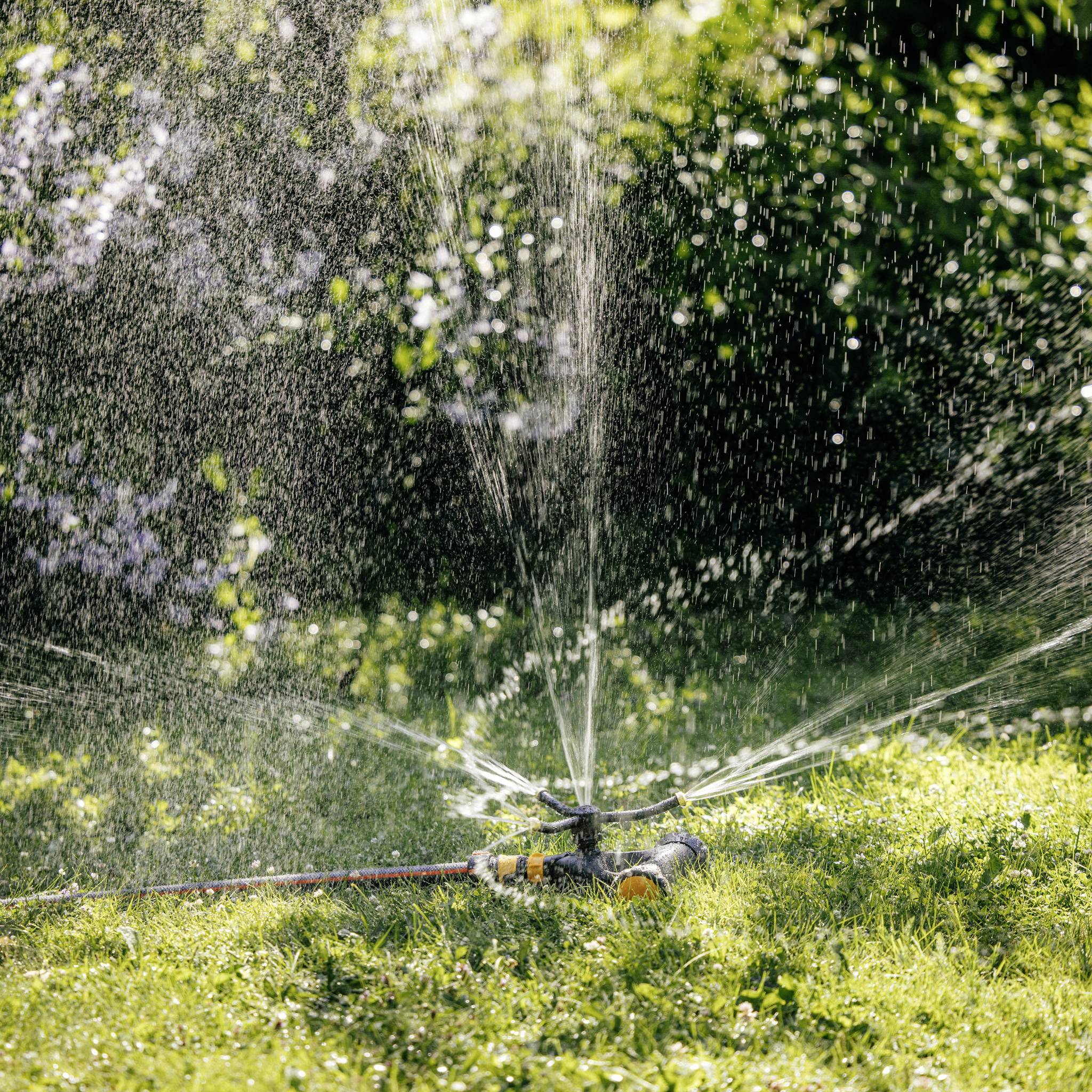 A lawn sprinkler is watering a green garden area in sunlight. Water droplets are sparkling in the air.