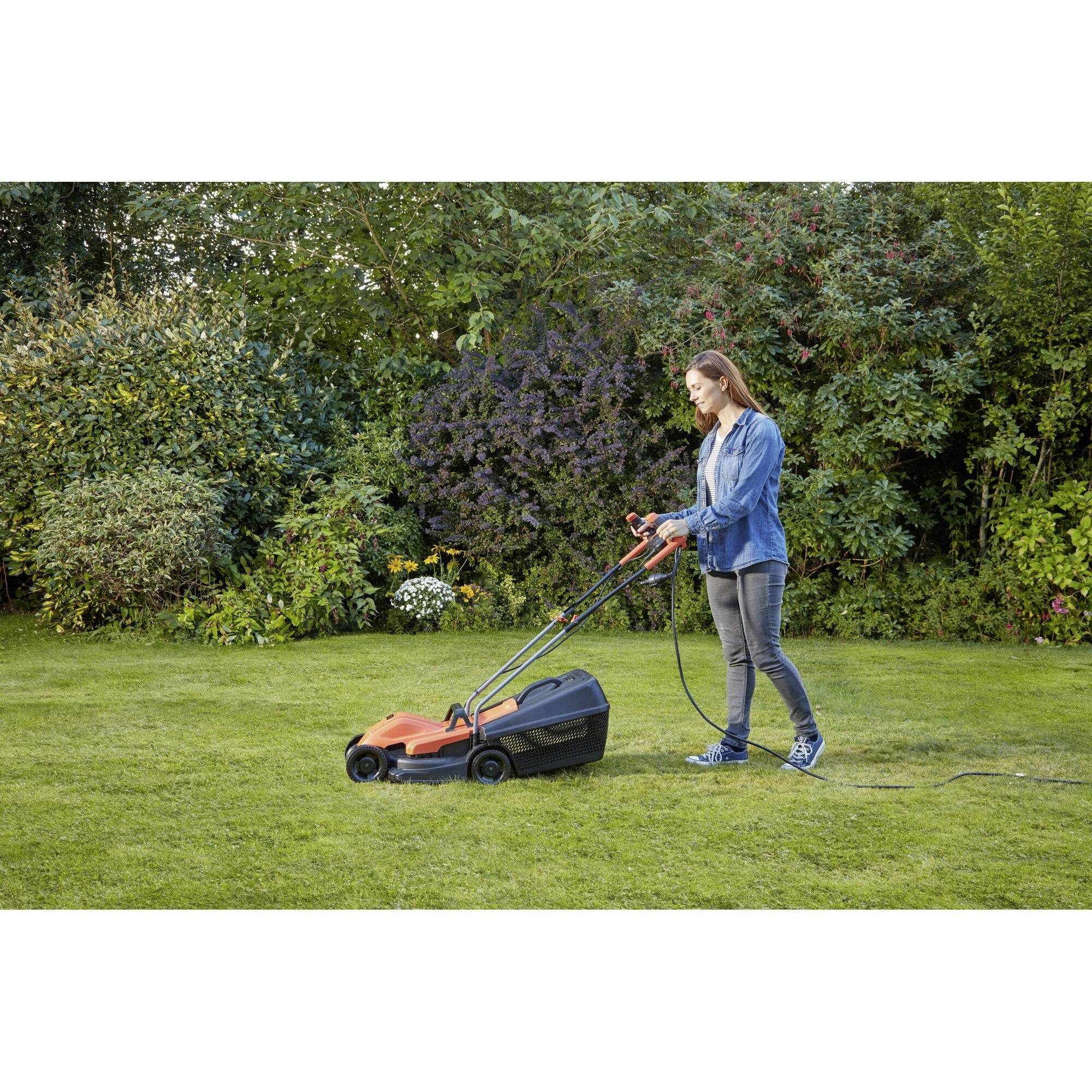 A person is mowing the lawn with an electric lawnmower in a garden with bushes and trees in the background.
