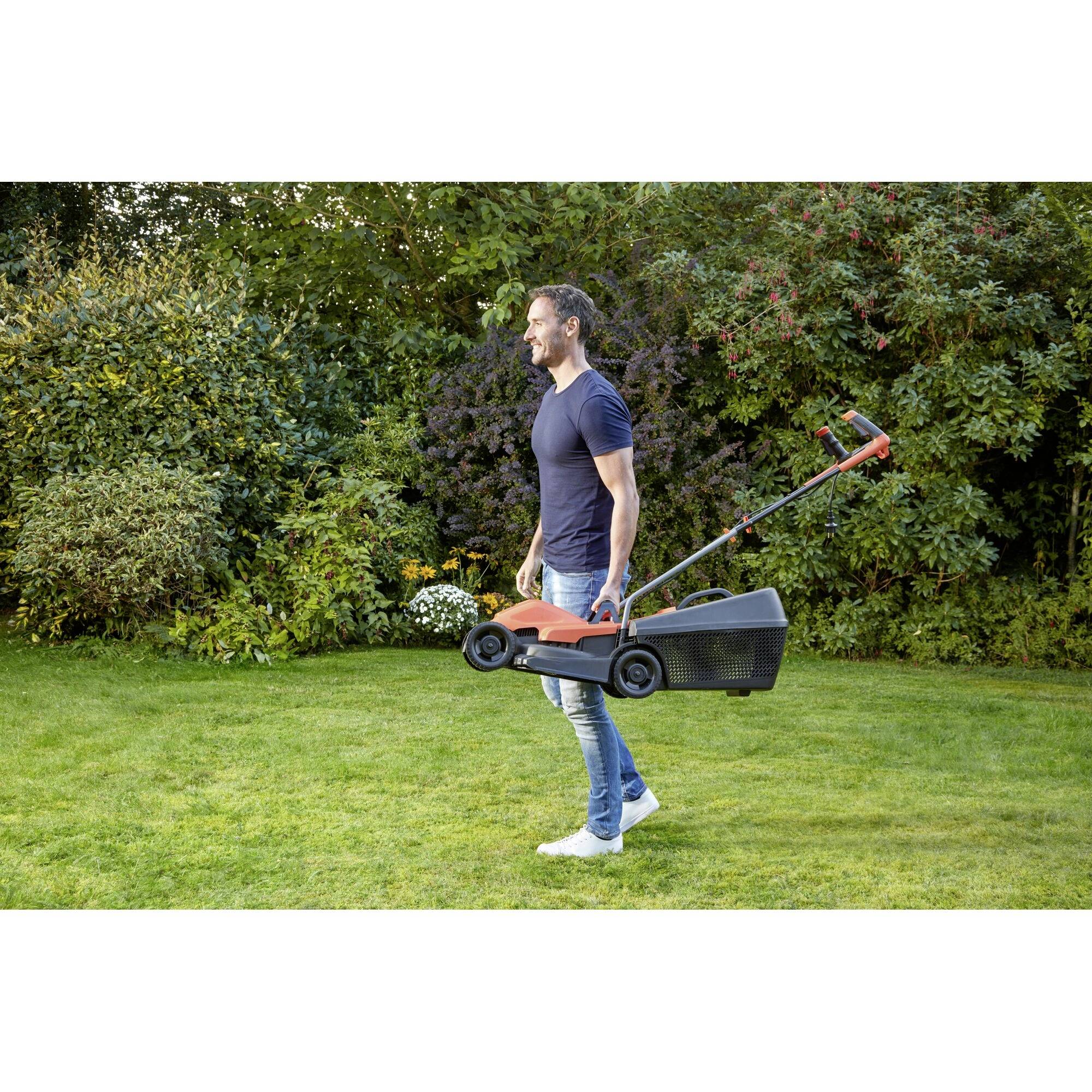 A man is carrying a lawnmower in a garden. The lawnmower is small and compact, suitable for straightforward garden maintenance.