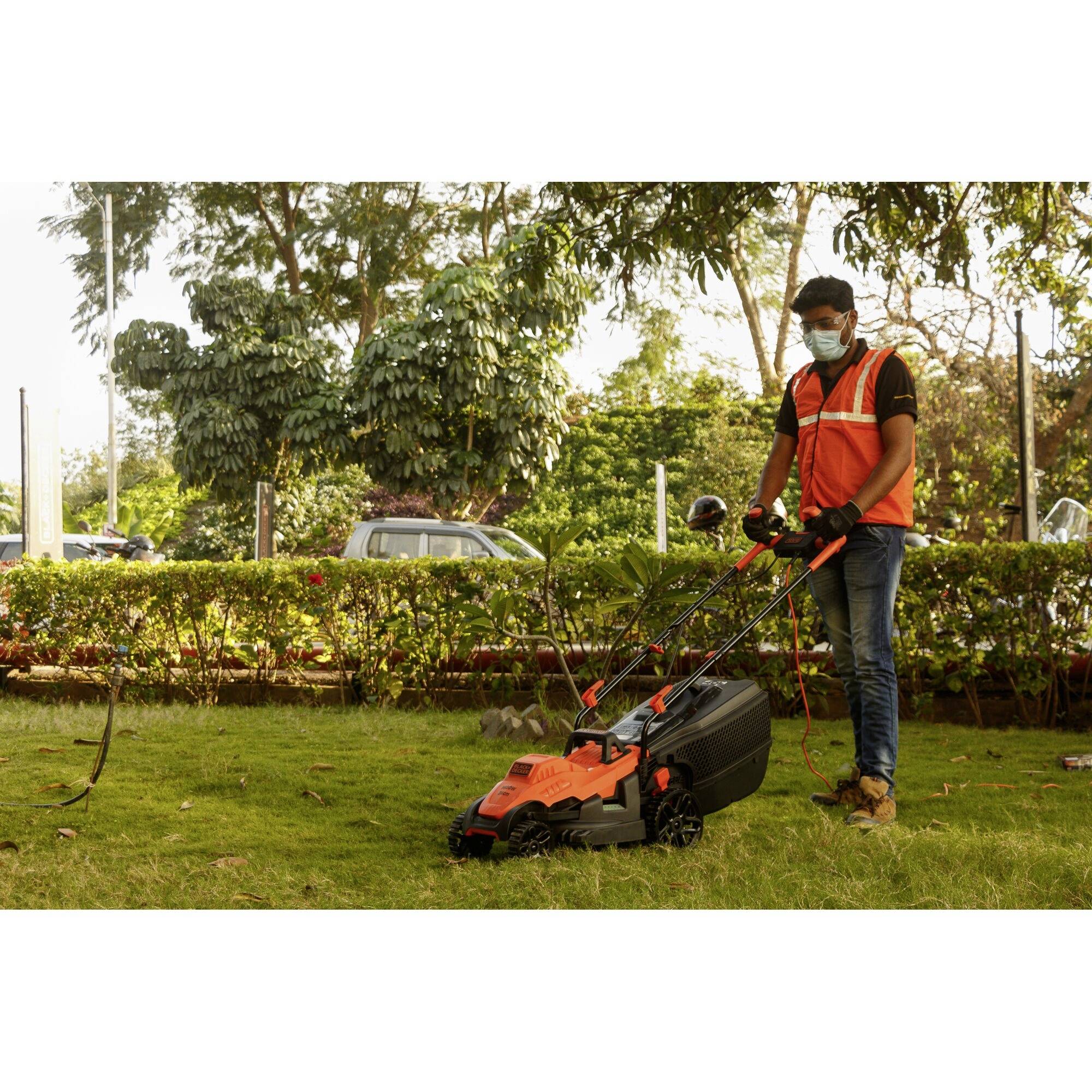 A person is mowing the lawn during the day in a garden with a lawnmower, wearing an orange hi-vis vest and a face mask.