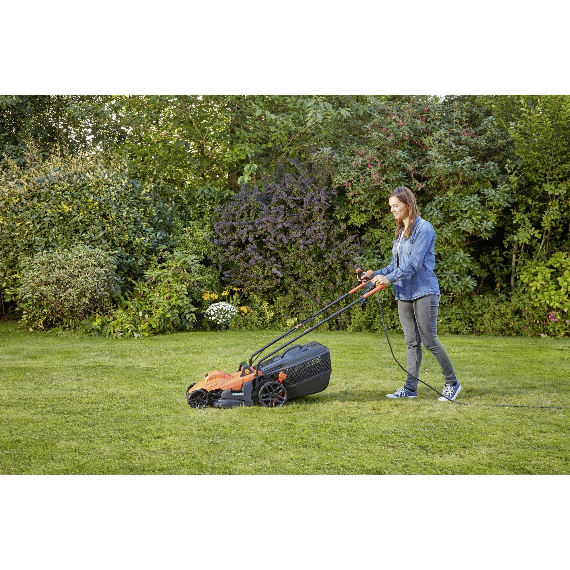 A woman is mowing the lawn with an electric lawnmower on a green lawn, surrounded by bushes and trees.