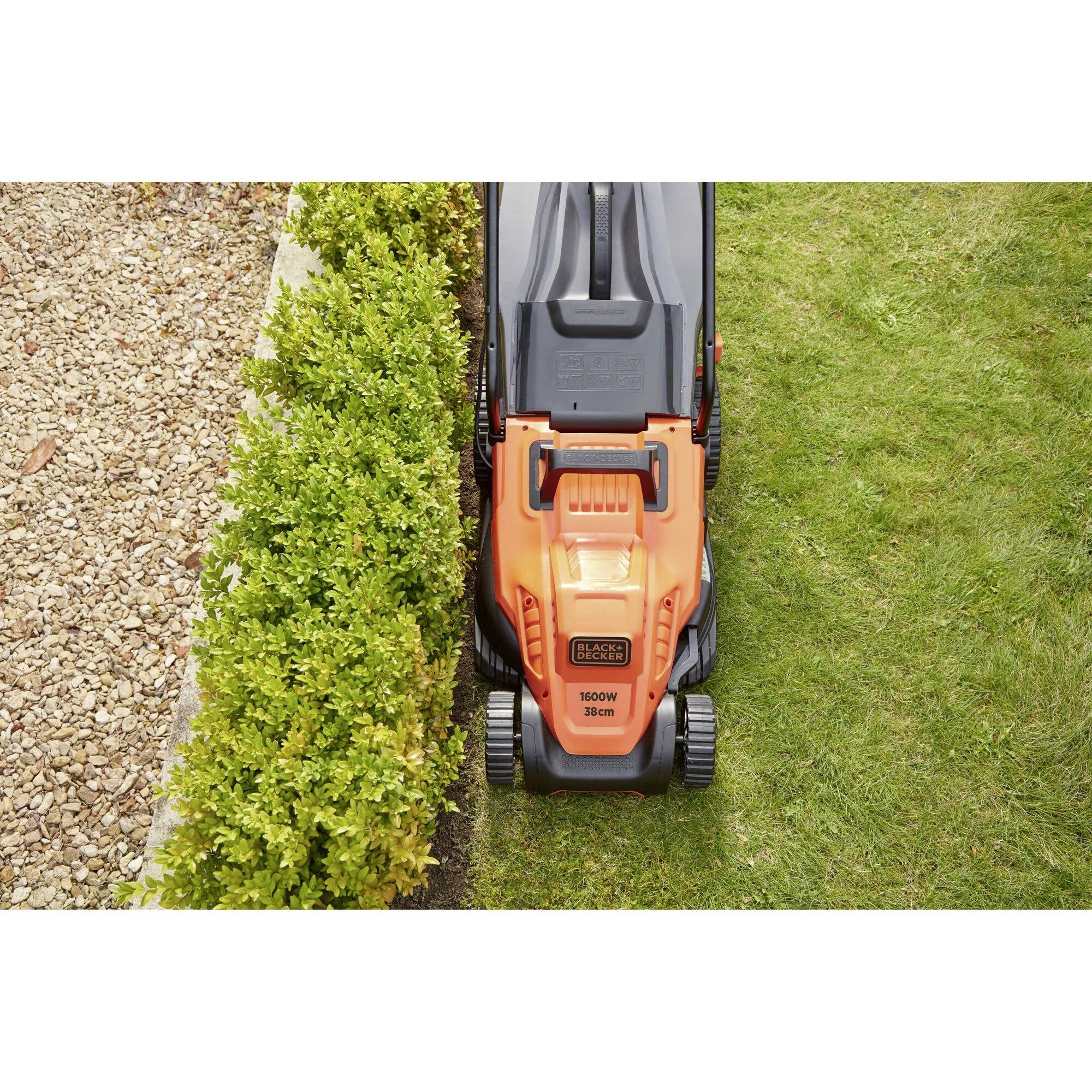 A lawnmower is cutting grass in a garden, with a hedge on the left and a gravel path on the right.