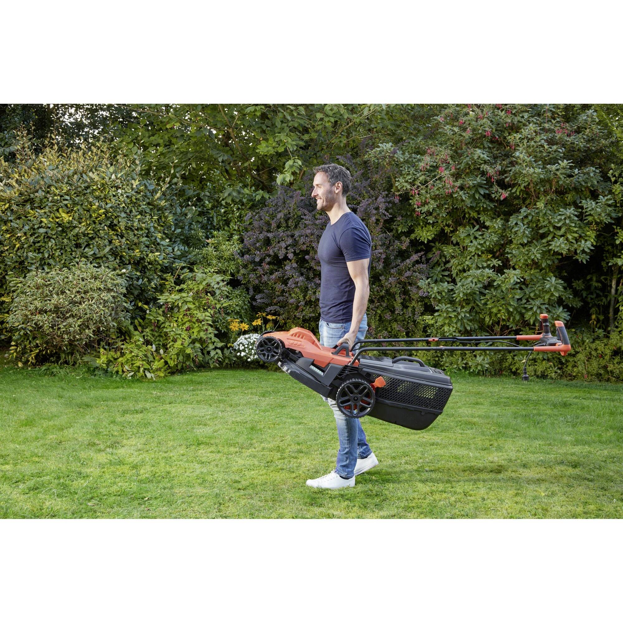 A person is carrying a lawnmower in a garden. Bushes and trees can be seen in the background.