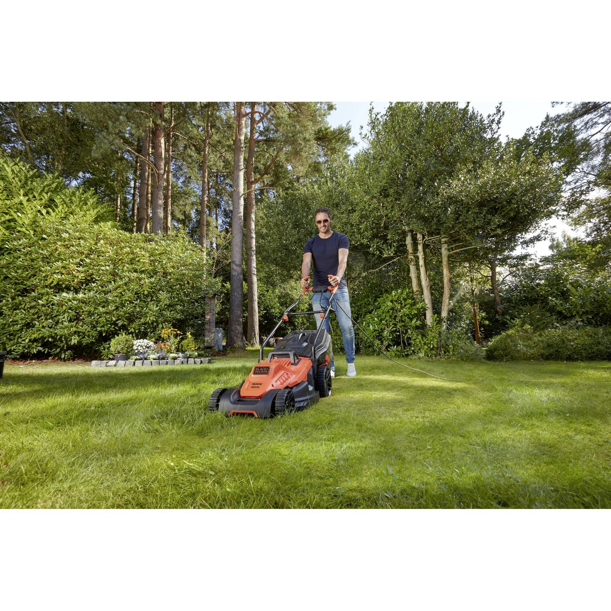 A person is mowing the lawn with an orange lawnmower in a sunny garden, surrounded by tall trees and shrubs.