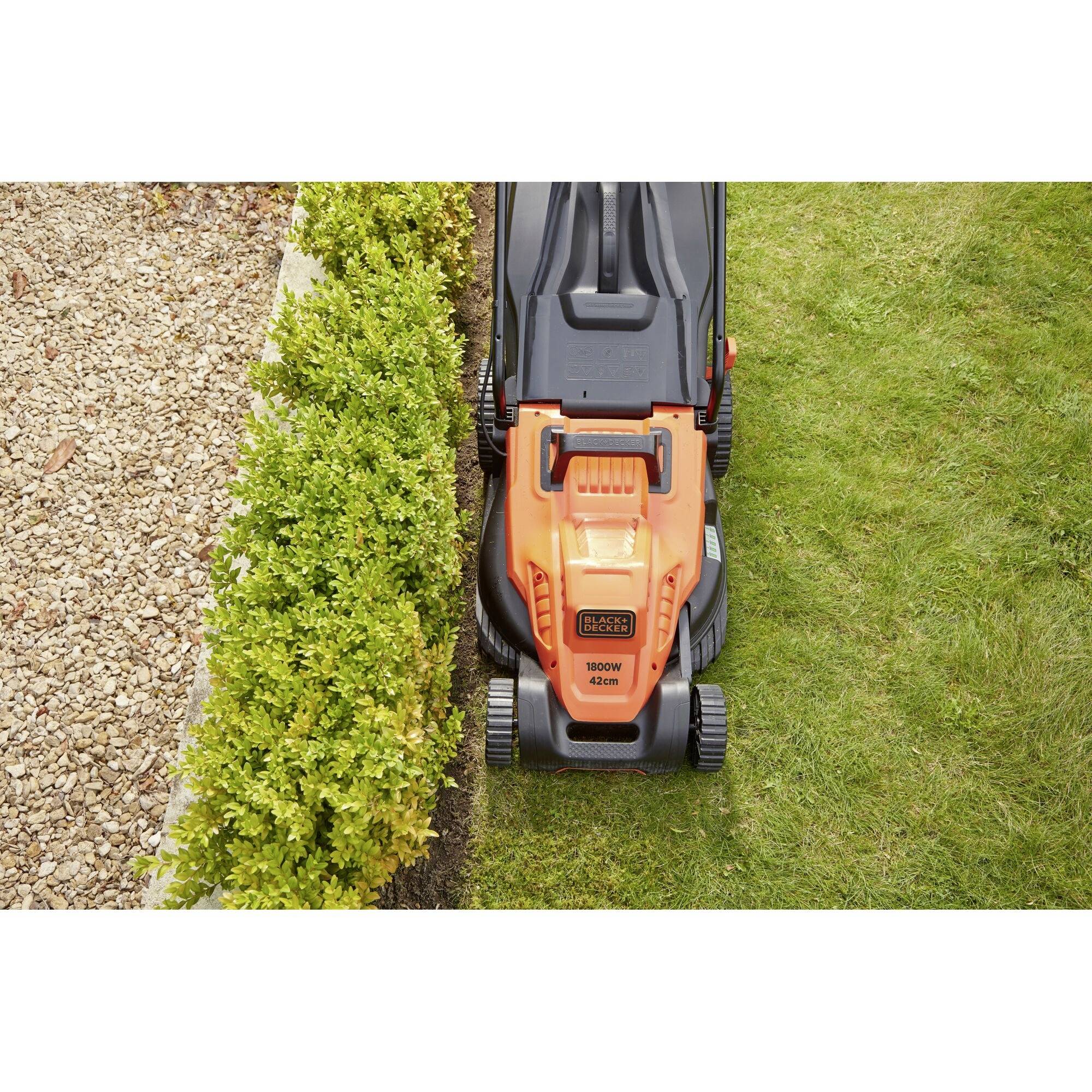'Lawnmower cuts grass beside a gravel path and a hedge. The lawnmower is orange and black.'
