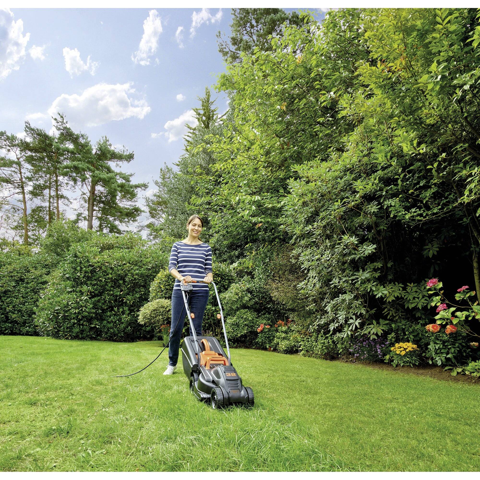 A person is mowing the lawn with an electric lawnmower on a sunny day in a green garden with trees and shrubs.