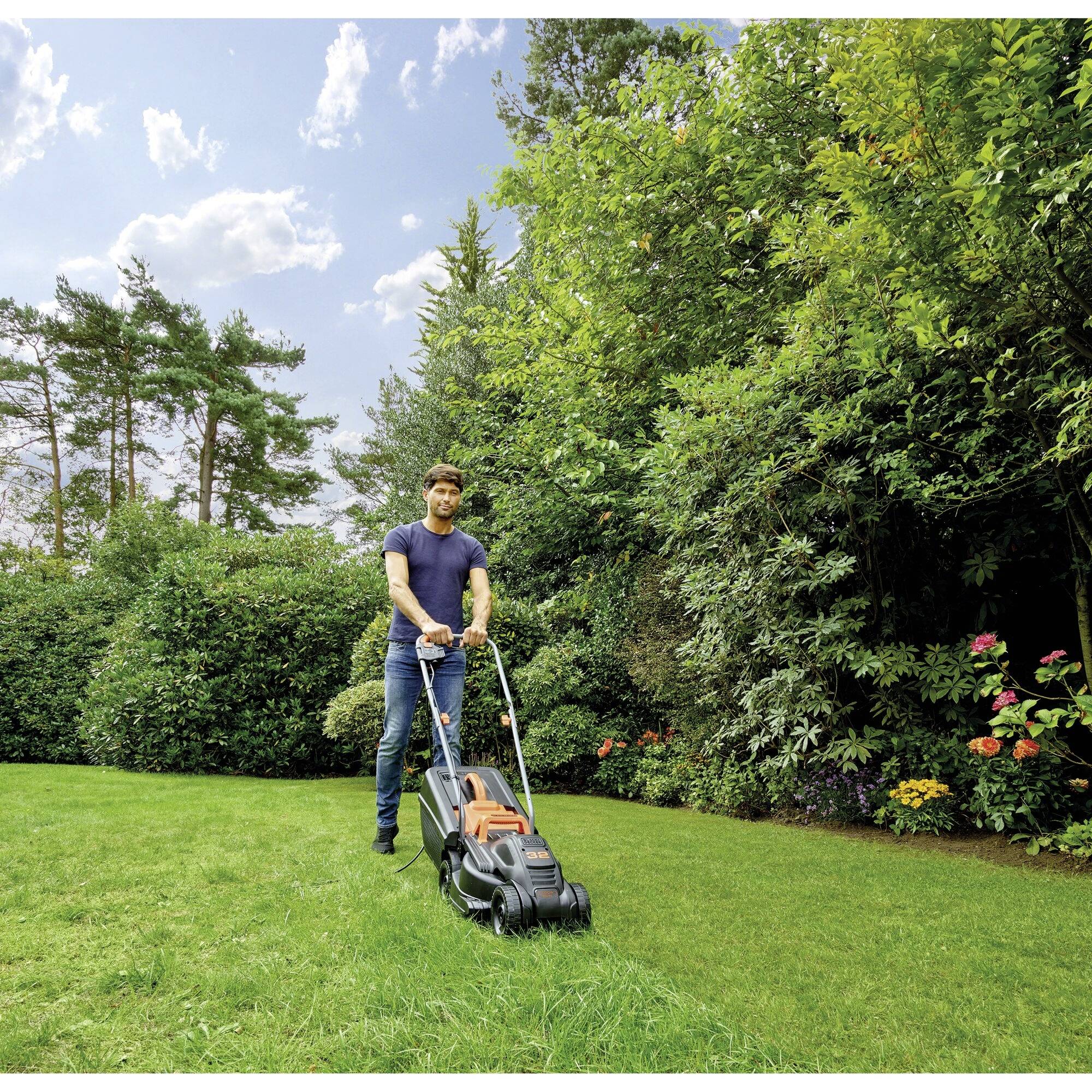 A person is mowing the lawn in a green garden with trees and flowering shrubs in the background.